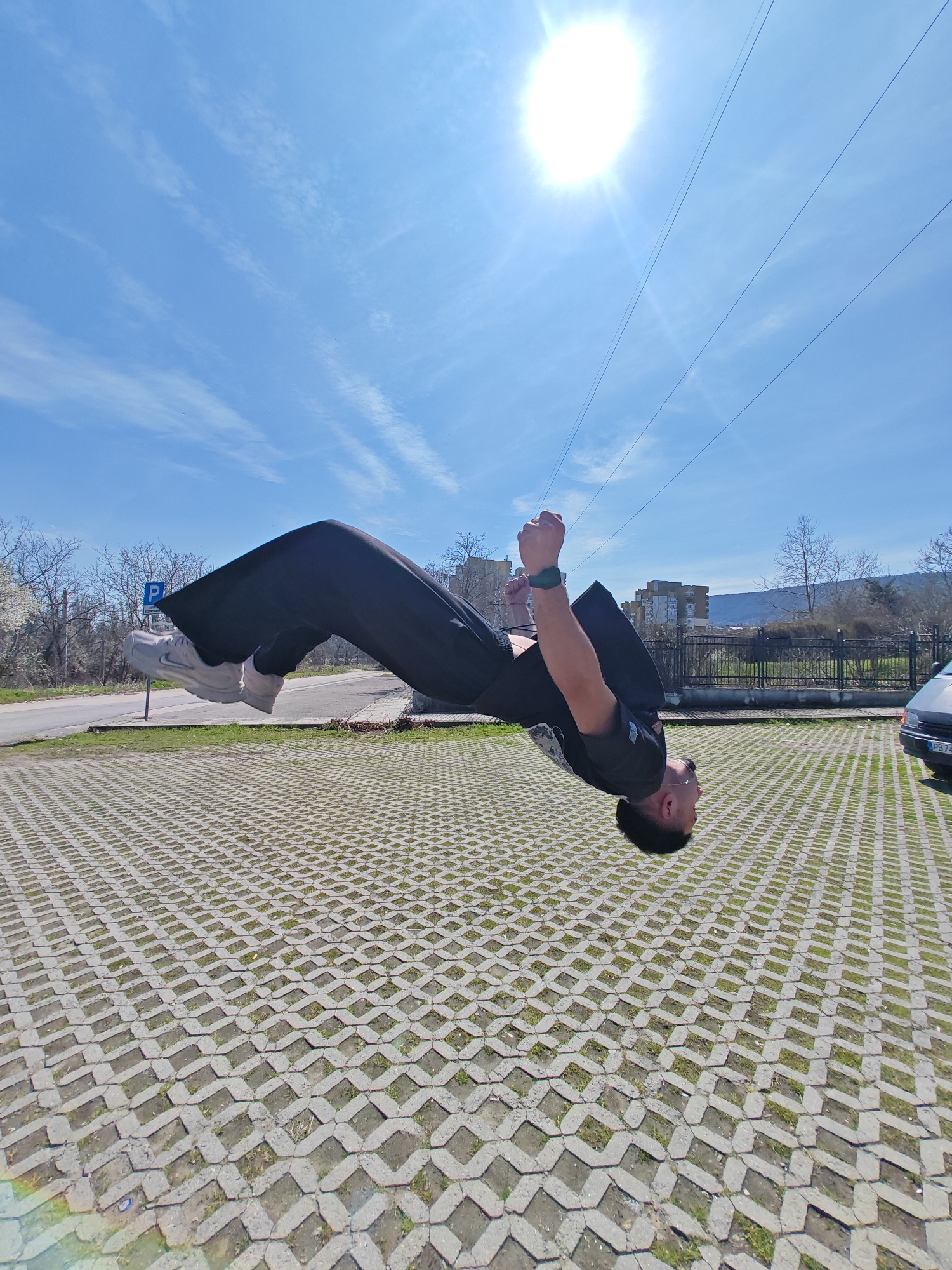 The image displays a male individual performing an aerial backflip maneuver outdoors in Shumen, Bulgaria. The subject is captured in mid-air, positioned horizontally and inverted with his head directed towards the lower-left. He is wearing a black short-sleeved t-shirt, black loose-fitting pants, and white athletic shoes; a dark green smartwatch is visible on his left wrist. His hands are clenched into fists near his chest. The ground surface consists of interlocking concrete pavers with green grass growing in the interstices, indicative of a parking area. In the background, from left to right, a paved road, a blue parking sign with a white 'P' symbol, and a black metal fence are visible. Beyond the fence, bare deciduous trees, multi-story residential buildings, and distant hills or mountains are present under a clear horizon. A silver vehicle is partially visible on the far right side of the frame. The sky is a vivid blue with scattered wispy clouds, and a brilliant, circular sun with prominent rays is positioned in the upper right quadrant. Several thin, dark utility lines traverse the upper portion of the sky. The scene is brightly illuminated by direct sunlight.