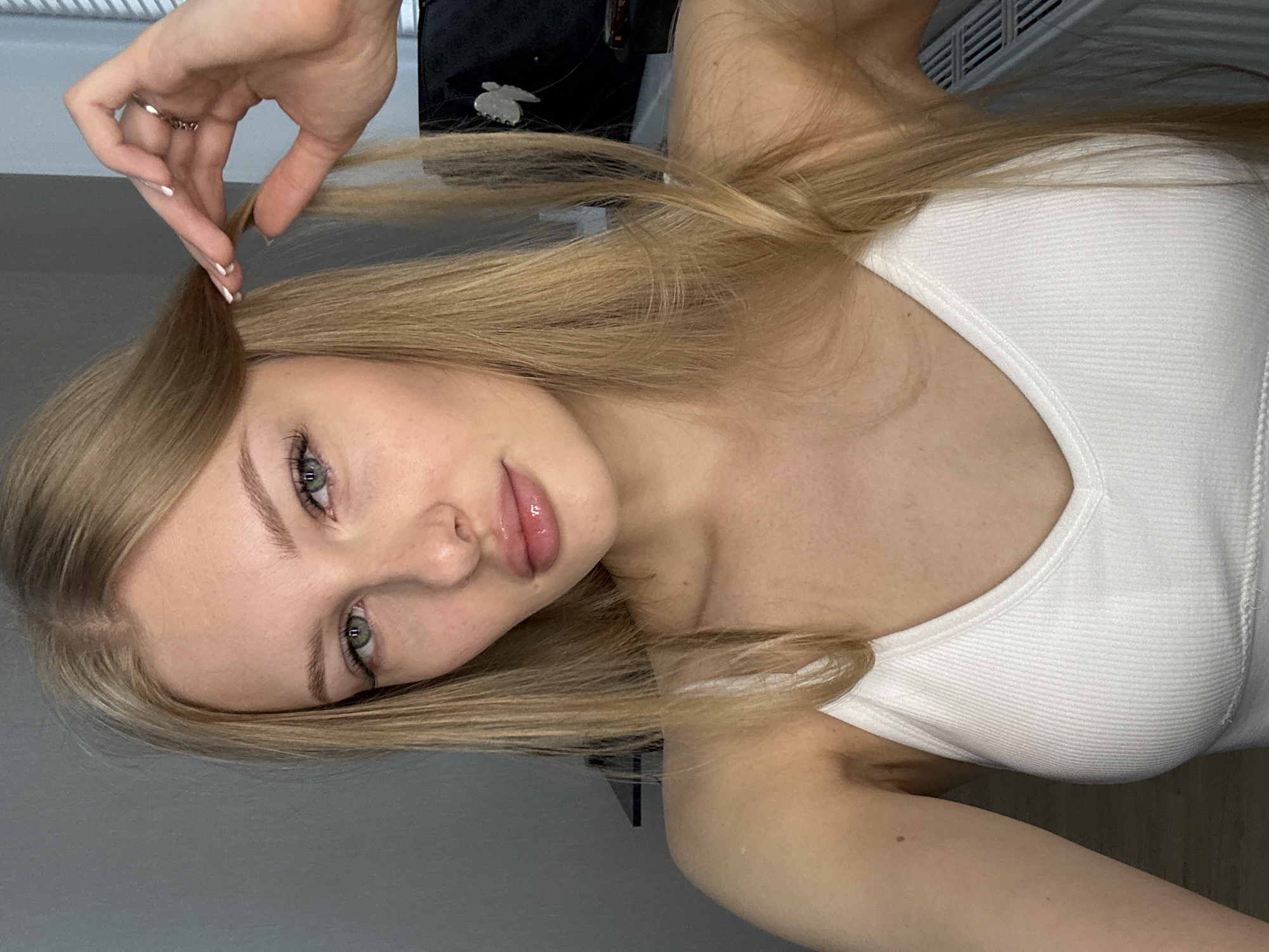 A young woman with blonde hair is shown in a close-up, self-taken photograph. She is wearing a white ribbed tank top. Her right hand is raised, touching her hair near her temple. She has light-colored eyes, styled eyebrows, and full lips with lip gloss. Her skin appears fair. The background is a plain, neutral wall. No specific location in Perm, Russia, is identifiable from the image.