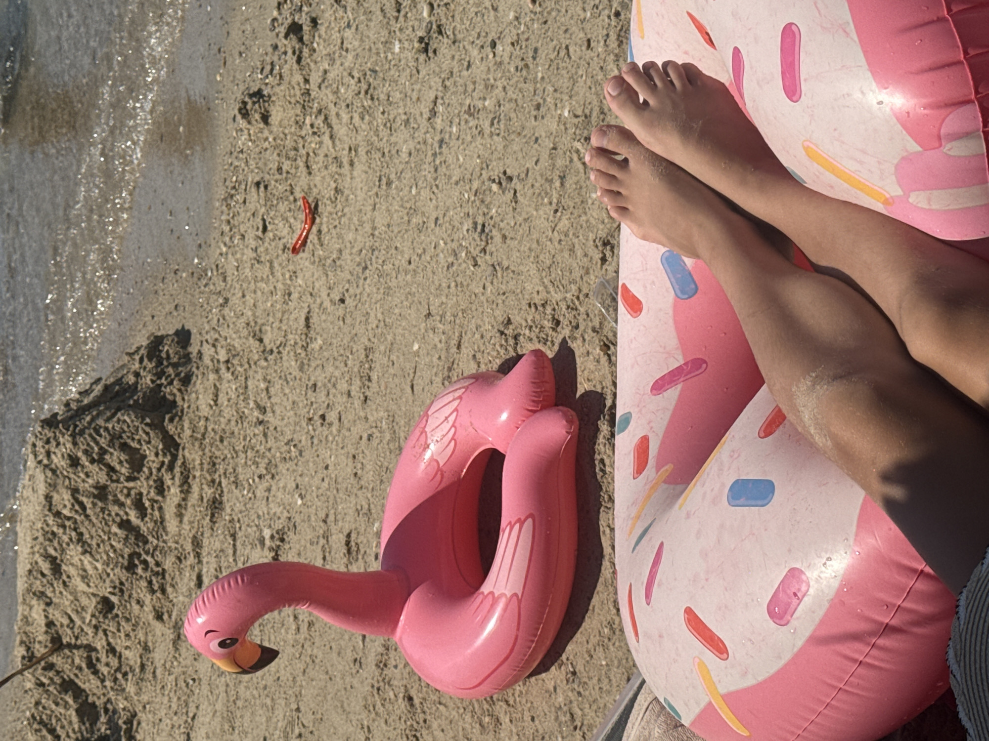 The image depicts a sunny day at a beach in Bodrum, Turkey. In the foreground, a person's bare legs and feet rest on a pink inflatable donut-shaped float adorned with colorful sprinkles. To the left of the legs and float, a pink inflatable flamingo float is partially visible, lying on the sand. The beach itself is composed of fine, light-colored sand with some small pebbles and shells. In the background, the calm turquoise waters of the Aegean Sea gently lap at the shoreline. A small pile of sand is also visible in the mid-ground. The scene suggests a relaxed atmosphere, typical of a vacation or leisure day by the sea.