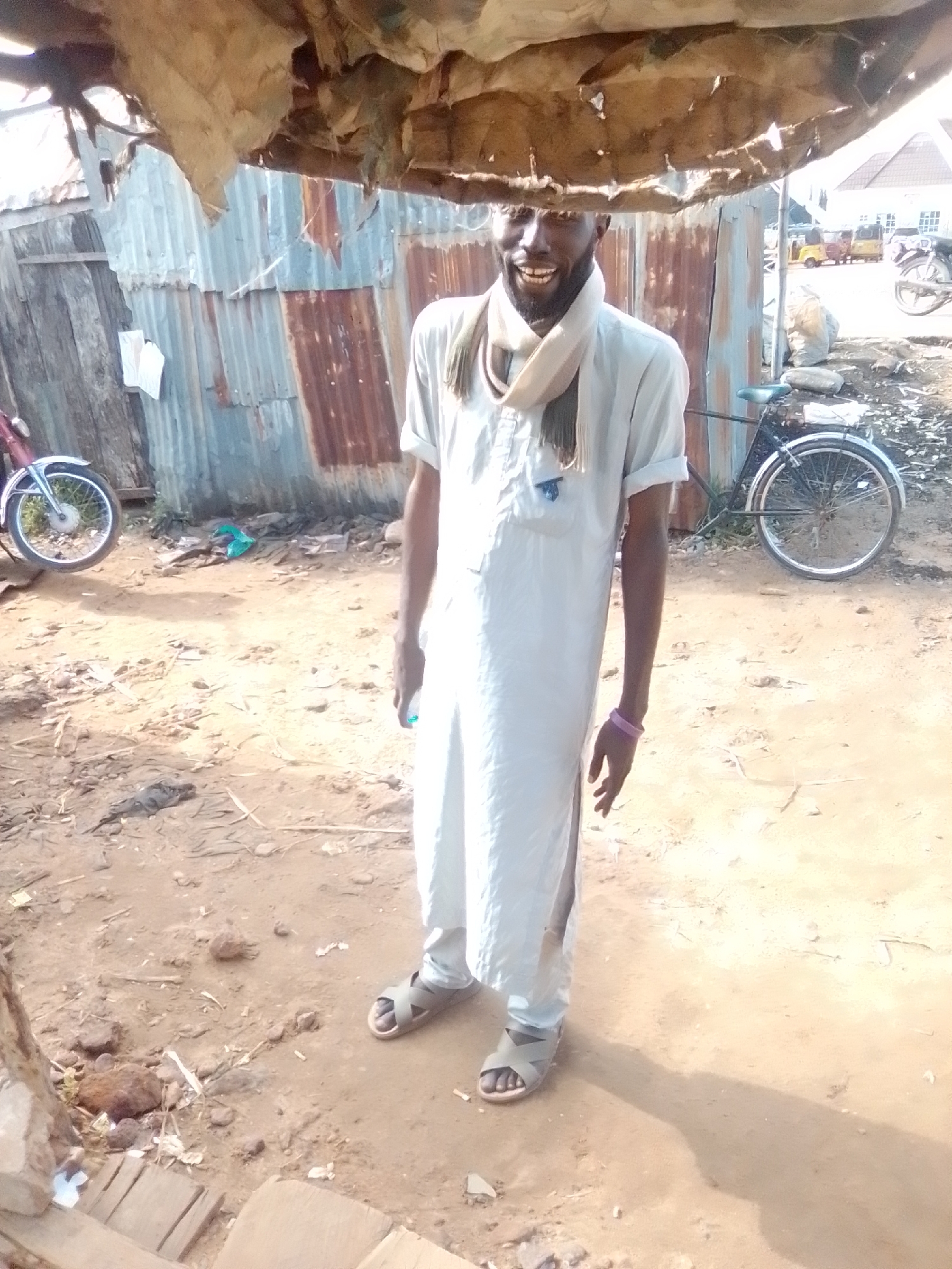 A medium shot captures a Black man standing in a dirt lot under a makeshift shelter in Pantami, Nigeria. He is wearing a light blue, ankle-length robe and a cream-colored scarf around his neck. His arms are bare, and he sports a purple bracelet on his left wrist. The man is smiling widely, looking towards the viewer. His sandals are brown and strapped. In the background, a corrugated metal wall is visible, along with a bicycle and what appear to be auto-rickshaws, suggesting a marketplace or bustling area. The ground is uneven, with scattered debris and small rocks. The shelter above is constructed from fabric and possibly wood. The overall scene depicts an outdoor, possibly informal, setting in a Nigerian city.