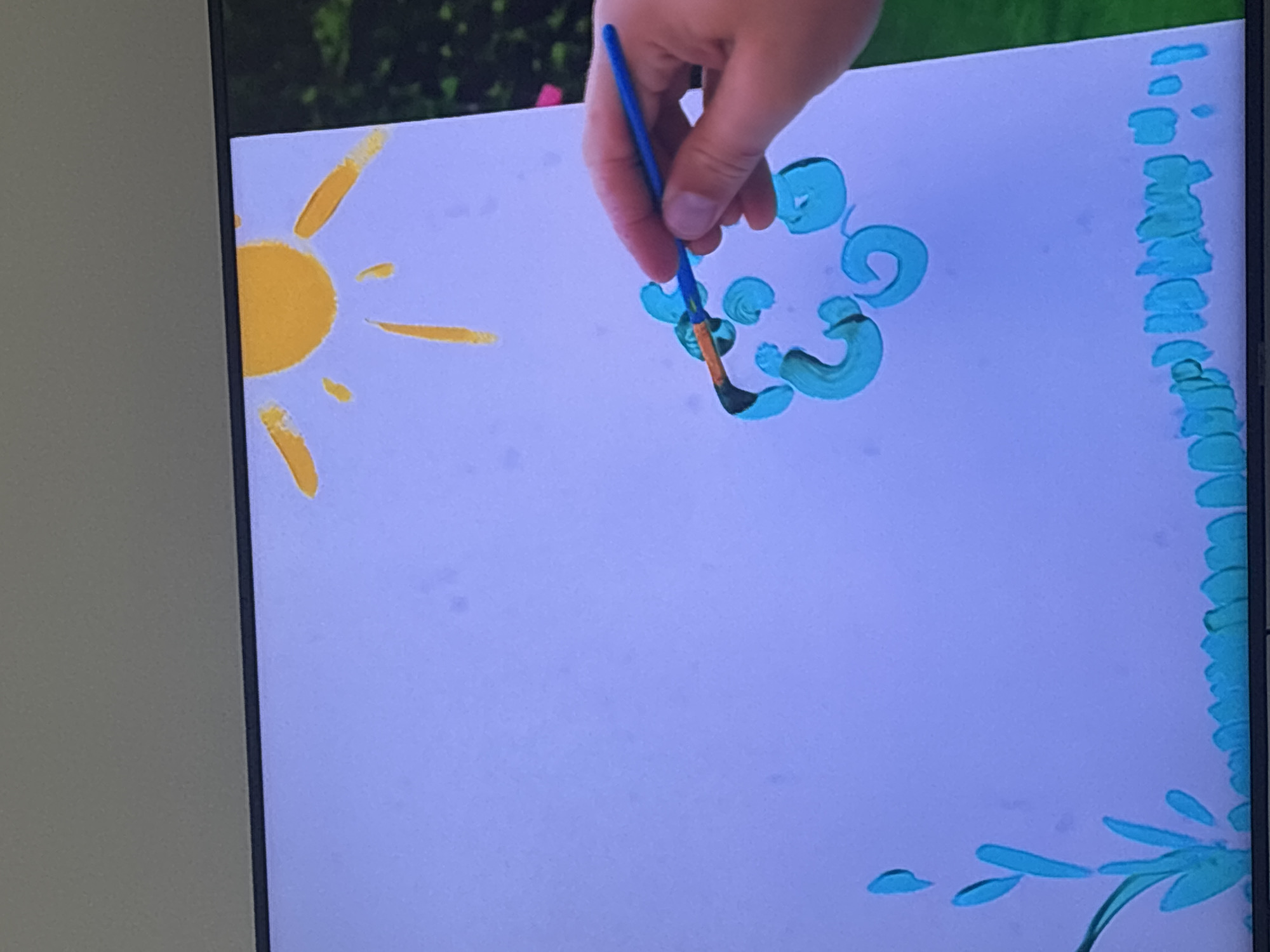 The image depicts a close-up view of a person's hand holding a blue paintbrush, actively applying teal paint to a white canvas. The painted elements visible on the canvas include a sun with yellow rays in the upper right corner, and abstract teal shapes that appear to be forming a cloud in the center. In the lower left and bottom of the canvas, there are teal strokes representing grass or plants. The background of the canvas is a light purple, and beyond the canvas, a blurred green and pink natural landscape is visible, suggesting an outdoor setting. The scene is shot from a slightly elevated, head-on perspective. There is no indication of the city of Dubai, United Arab Emirates.