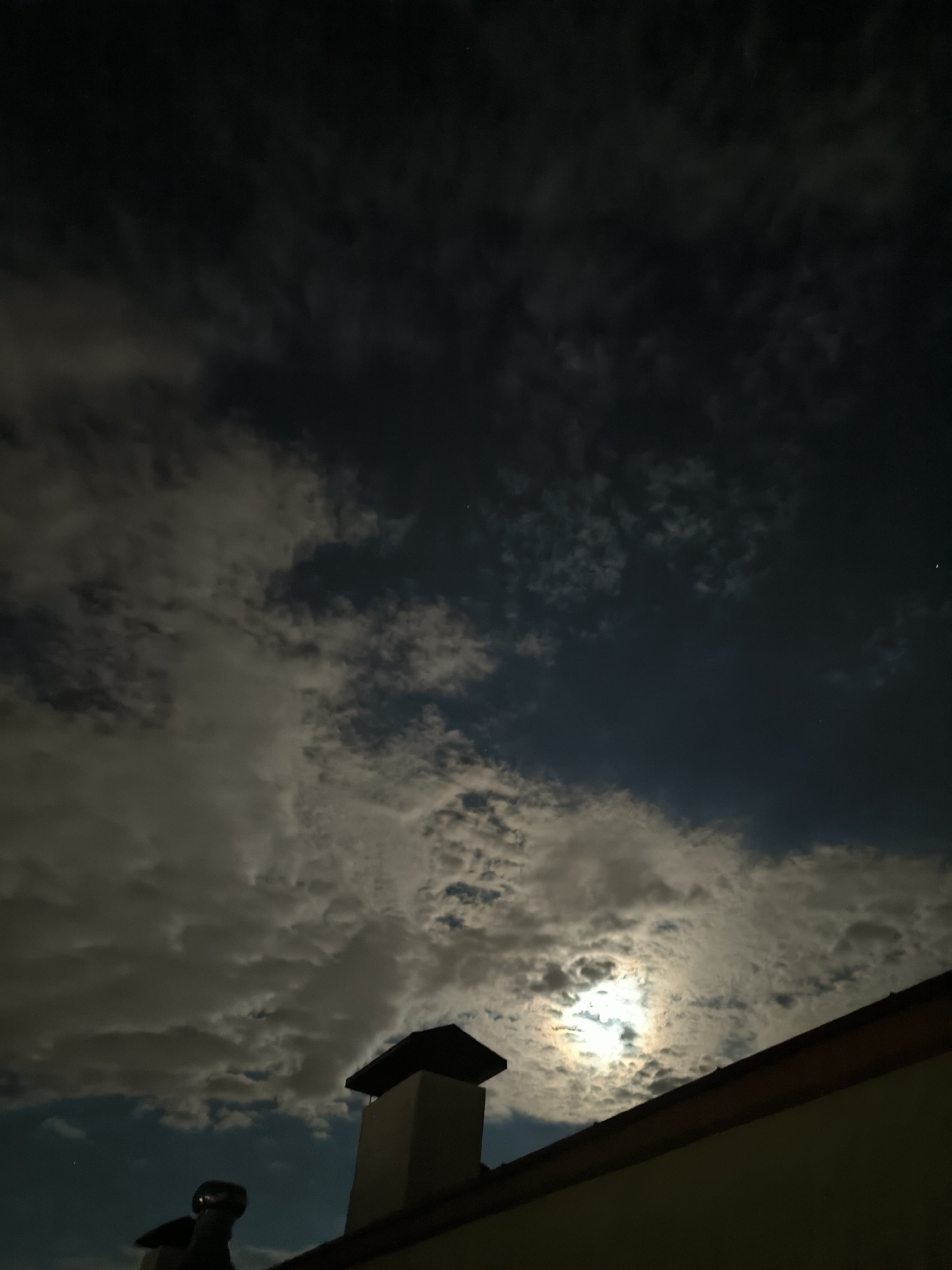 The image captures a night sky over Chișinău, Moldova, with a full moon partially obscured by clouds. The moon's bright light illuminates the edges of some of the clouds, creating a dramatic effect. In the foreground, the silhouette of a rooftop is visible, with a chimney and another smaller structure, possibly a vent pipe. The clouds vary in density, with some dark, heavy masses and others thin and wispy, allowing the moonlight to shine through. A few faint stars are visible in the darker patches of the sky. The scene is serene and atmospheric, suggesting a quiet night in the city.