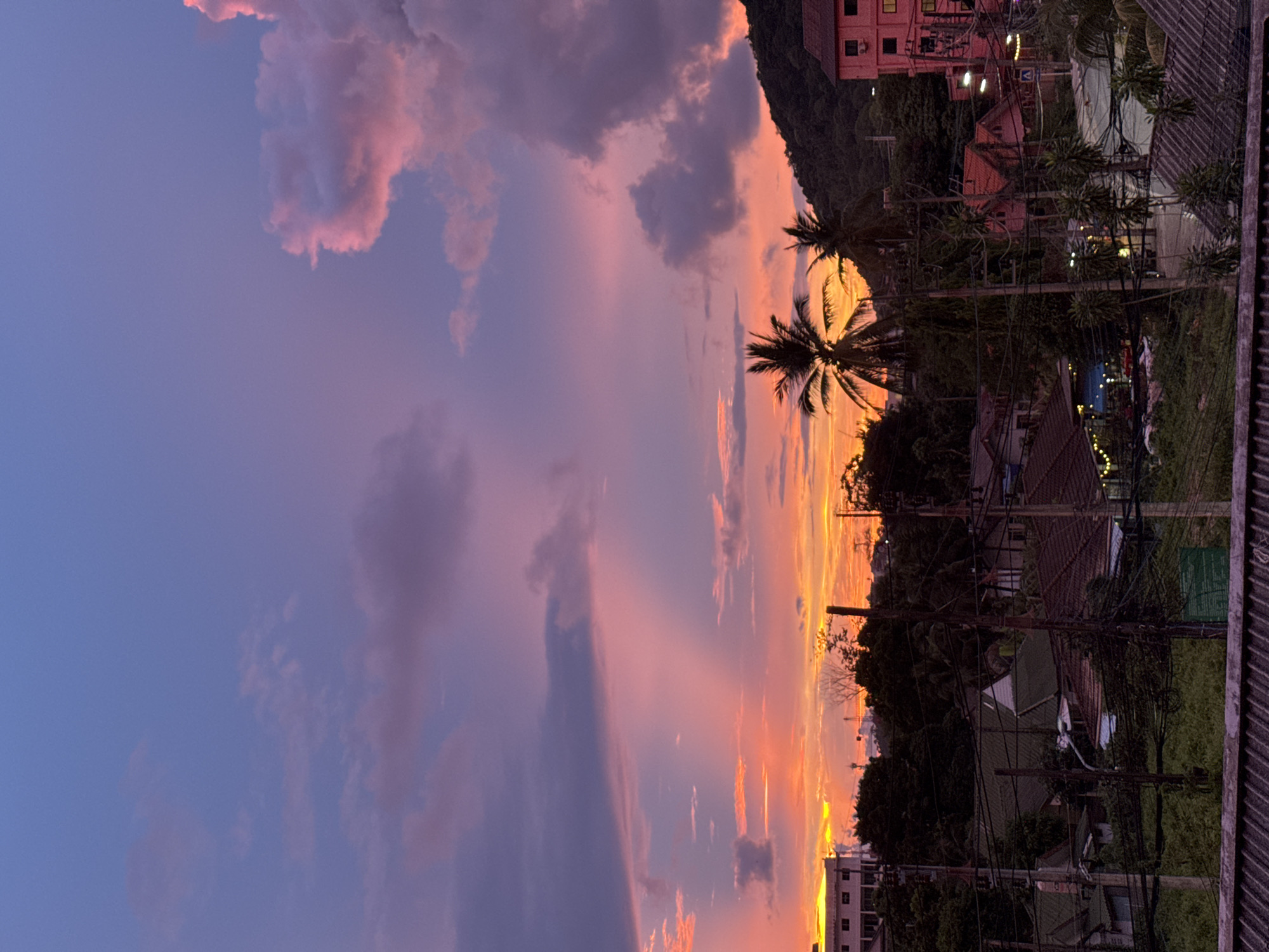 The image depicts a tropical sunset scene in Karon, Thailand. The sky is filled with vibrant hues of pink, purple, and orange, indicating the golden hour. Wispy clouds are scattered across the sky, catching the warm light of the setting sun.
In the foreground, palm trees with their characteristic fronds are silhouetted against the colorful sky. Beyond the trees, a collection of buildings and houses can be seen, suggesting a residential or commercial area. The architecture appears to be in a tropical style, with tiled roofs. Electrical wires and poles are visible, strung between the buildings, common in many urban and semi-urban tropical landscapes.
The overall atmosphere of the image is serene and tranquil, characteristic of a sunset. The warm colors of the sky evoke a sense of peace and beauty. The location cues, such as the palm trees and the style of buildings, along with the provided location context of Karon, Thailand, strongly suggest this is a scene from a coastal town in Southeast Asia. There is no visible text in the image. The time of day is clearly sunset, with the weather appearing clear and pleasant, allowing for the dramatic sky colors.