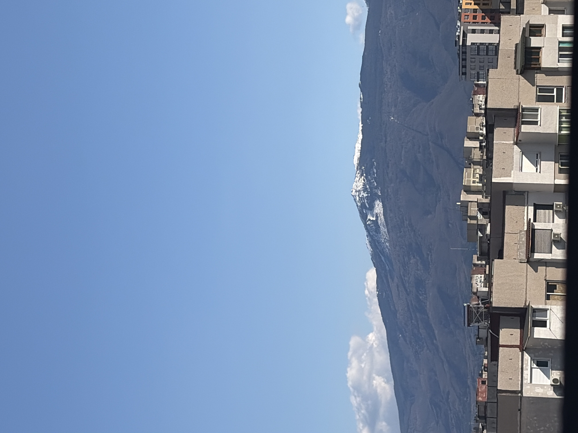 The image shows a mountain range and buildings against a clear blue sky. The buildings appear to be apartment blocks, typical of urban residential areas. The mountain range is located next to the apartment blocks. The image shows a location in the city of Sofia, Bulgaria where you can see the mountain range.