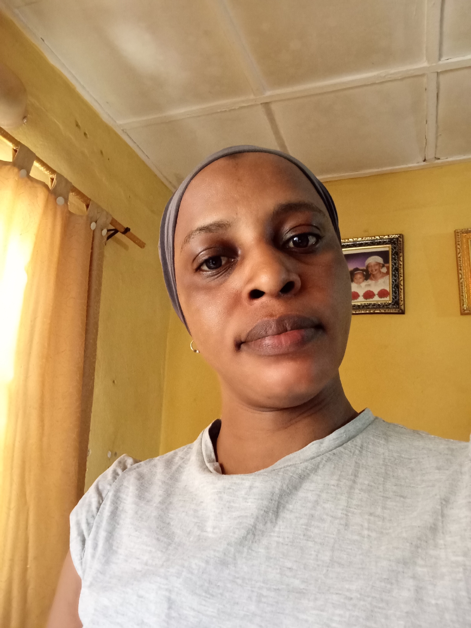 The image shows a woman with dark skin, wearing a gray head covering and a gray t-shirt. She is looking directly at the camera. She has short hair covered by the headscarf. Behind her is a yellow wall with two framed pictures. A yellow curtain is visible to the left. The location is identified as Narayi, Nigeria.