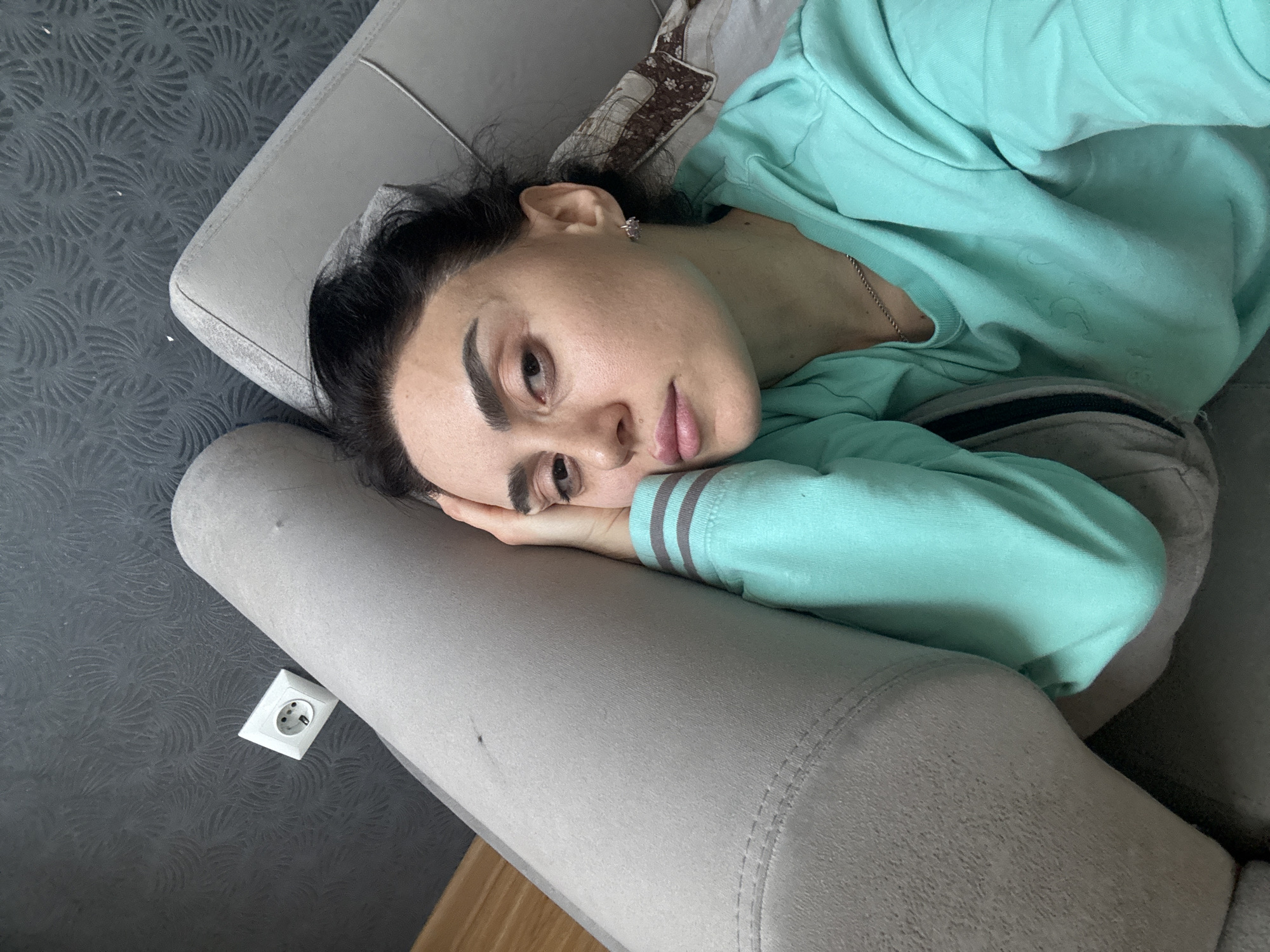 A woman is lying on a gray couch. She is wearing a light blue long-sleeved shirt. Her head is resting on her hand. An electrical outlet is visible on the wall. The exact location in Tbilisi, Georgia, cannot be determined from the image.