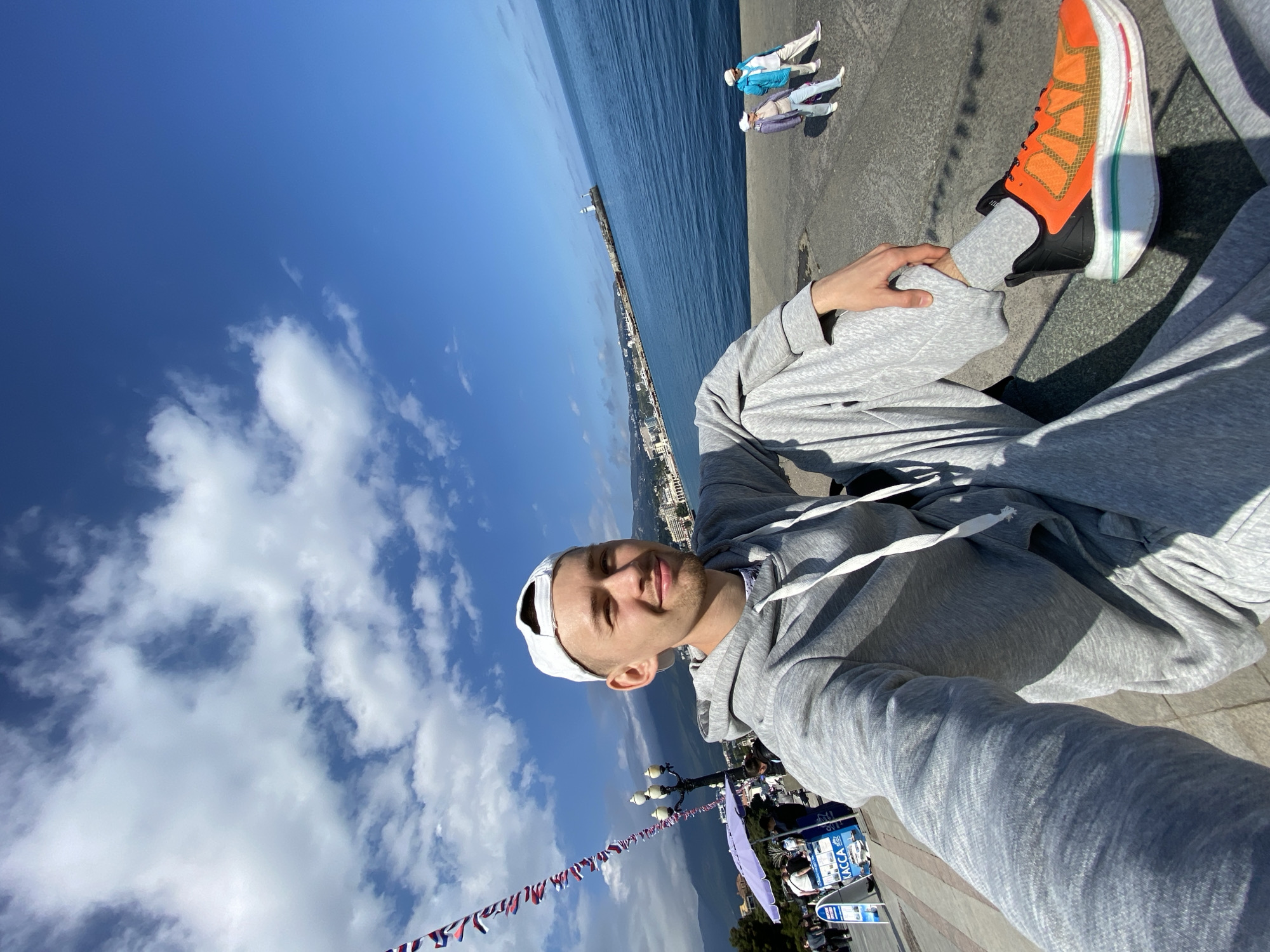 A man is sitting on a ledge in what appears to be the city of Yalta, Ukraine. The image is taken from a low angle, showing the man from the waist up. He is wearing a grey tracksuit, a white cap, and orange and black sneakers. The man is holding his leg up.
In the background, there is the sea, a strip of the city along the coastline, and a blue sky with white clouds. The ledge appears to be part of a promenade or walkway. A string of colorful flags is stretched along the ledge near the man. Other people are visible in the background. There are what appears to be ticket kiosks and people sitting on benches.
