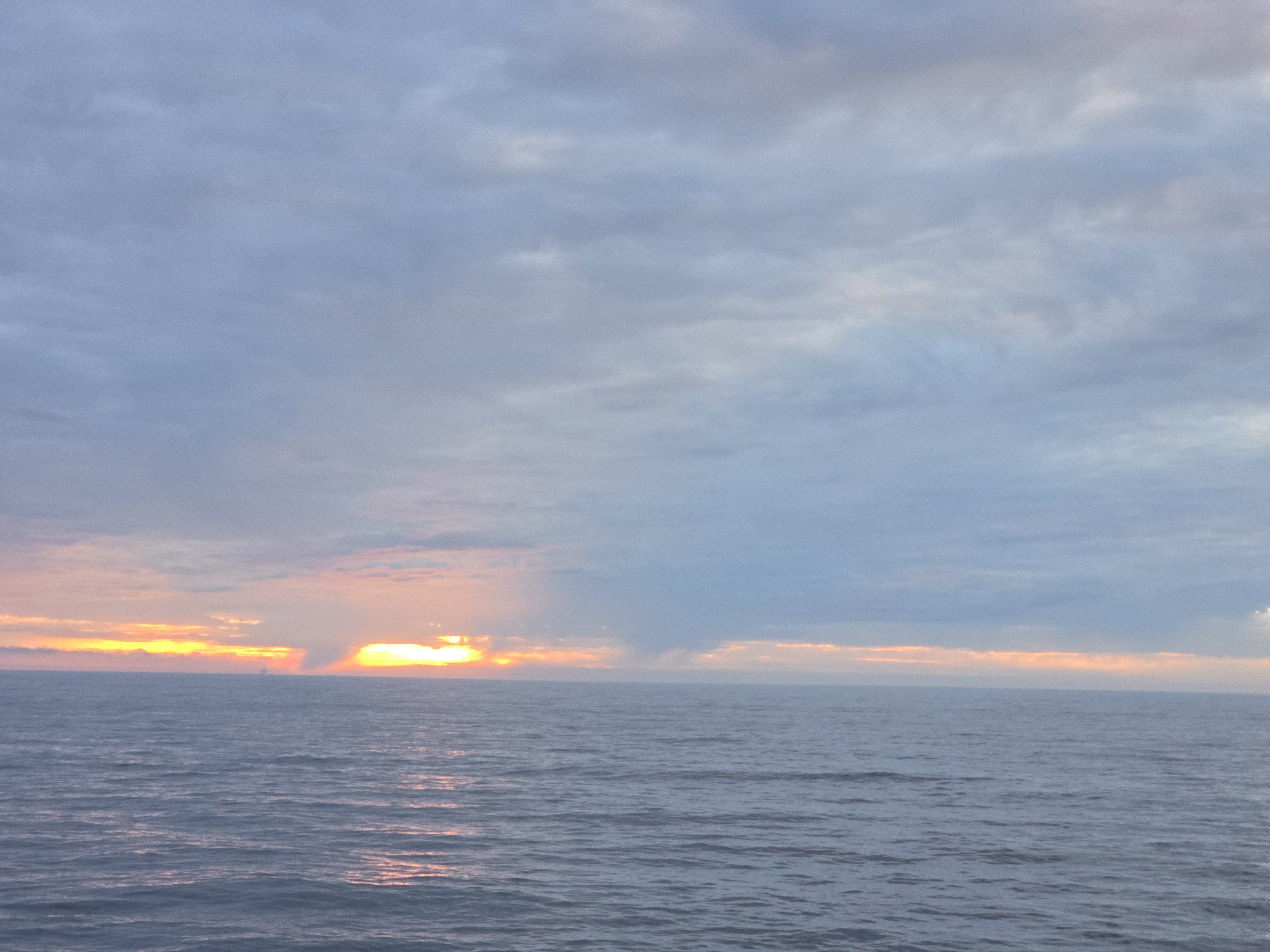 The image depicts a seascape at sunset. The sky is covered in cloudy formations ranging in color from blue to gray with hints of pink and orange near the horizon. The sun is setting on the horizon, casting an orange and golden glow on the water. The sea is calm, with gentle waves reflecting the colors of the sunset. No people, objects, or specific locations in the city of Unknown, Unknown, are discernible in this image.