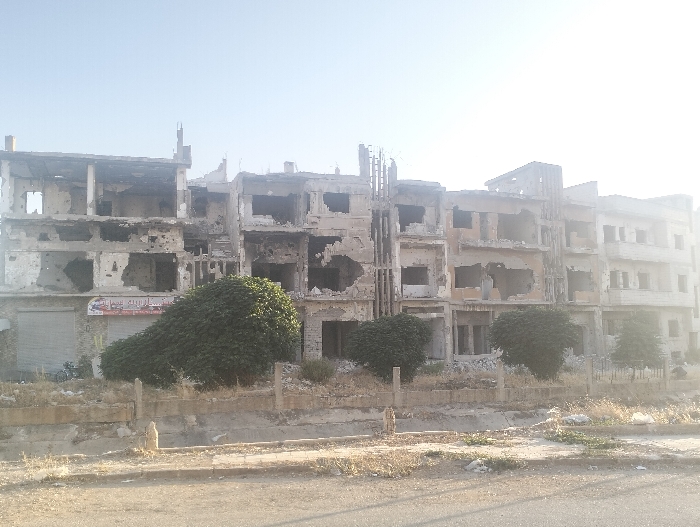 The image depicts a row of heavily damaged, multi-story concrete buildings in the city of Homs, Syria, under a clear, bright sky. The structures, likely former residential or commercial properties, exhibit widespread destruction characterized by large, irregular openings where windows and sections of exterior walls once stood. Numerous pockmarks and holes are visible across the facades, indicative of blast or impact damage.
The leftmost building features a grey metal roll-down security shutter on its ground floor, partially lowered, with a red and white sign above it displaying Arabic text, possibly advertising clothing. Its upper floors show significant structural compromise and burn marks. Adjacent buildings to the right display similar devastation, with exposed rebar and concrete slabs visible within their hollowed-out interiors.
In the foreground, a stretch of dry, unpaved ground with sparse, light-brown vegetation and scattered debris is present. A low, eroded concrete barrier or wall runs horizontally, separating the immediate foreground from the base of the damaged buildings. Several dense, dark green bushes, one notably large and pear-shaped, are situated between this barrier and the building facades. No individuals are visible in the scene.