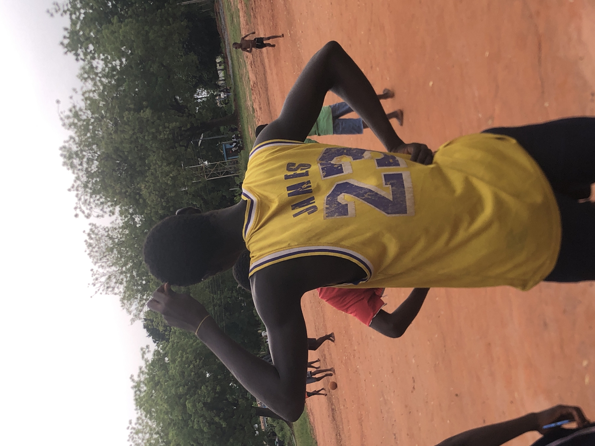 The image, oriented with the sky at the top, captures an outdoor scene in Nsugbe, Nigeria, likely on a field of reddish-orange laterite soil. The main subject is a person seen from behind in the foreground, wearing a vibrant yellow basketball jersey with "JAMES" and the number "23" printed in purple with a white outline on the back. This person has dark skin and is wearing dark shorts. Their left hand rests on their lower back, while their right arm is bent with the hand near their head.
In the mid-ground and background, several other individuals are visible on the dirt field, suggesting an informal gathering or sports activity. One person wears a green shirt and dark shorts, while another further back appears to be running or walking, clad only in shorts. A small, orange ball, possibly a basketball, is visible on the ground near some feet, indicating a game might be in progress. Lush green trees line the background and left side of the frame, under a bright, overcast sky. Some metallic structures, possibly scaffolding or parts of a stand, are partially visible beneath the trees. In the bottom right corner, a hand holding a smartphone is also visible. The setting appears casual, likely taking place during daylight hours.