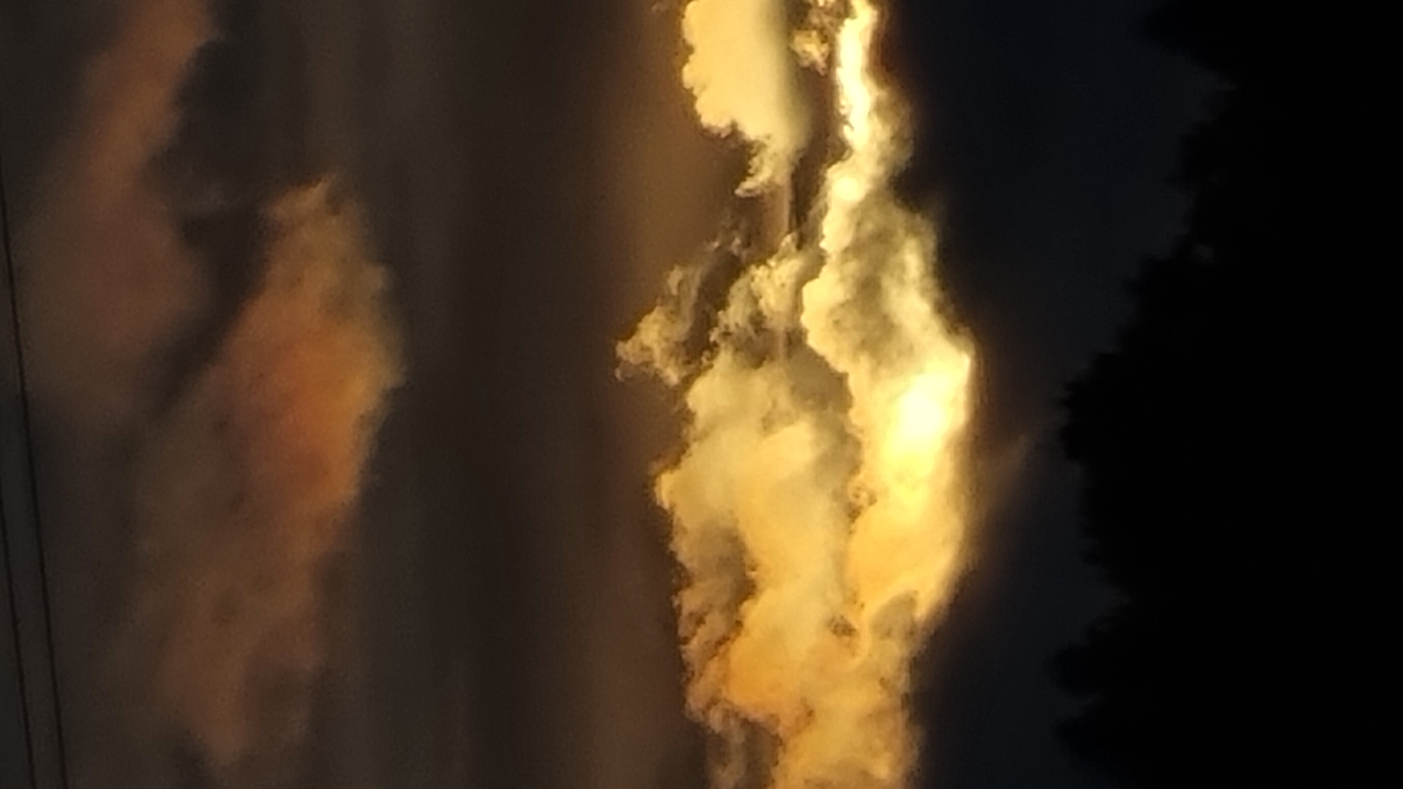 A horizontal, full-frame, eye-level shot captures a dramatic sky scene above a dark silhouetted treeline, possibly near Vysokoe, Russia. The sky is filled with thick, dark clouds tinged with orange and yellow hues, suggesting sunset or sunrise. A bright band of golden light illuminates the clouds from behind, creating a sense of depth and atmospheric drama. The foreground is dominated by the dark silhouettes of trees, which obscure any ground detail. The overall mood is one of natural beauty and a touch of mystery.