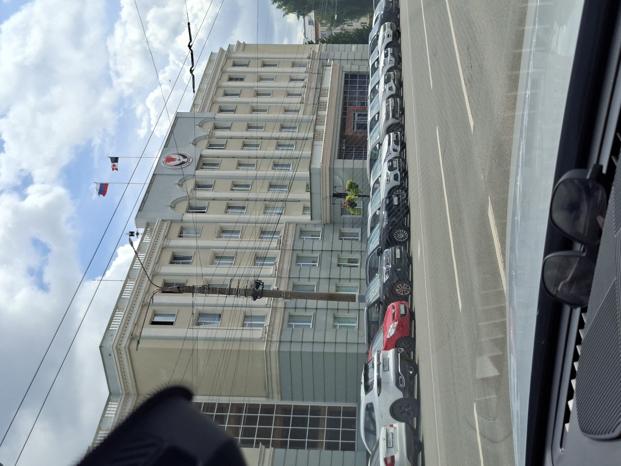 The image depicts a street scene in Izhevsk, Russia, with a large, imposing building serving as the primary subject. The building, which appears to be a government or administrative structure, features a light yellow facade with white architectural details and a light grey lower level. Several flags are visible on the roof, including the Russian flag and another flag with a black and red design. A prominent circular emblem is also displayed on the building's facade.
In front of the building, a row of parked cars lines the street. These include various makes and models, such as a white SUV, a red hatchback, and several dark-colored sedans. The cars are parked parallel to the sidewalk. The street itself is paved, with white lane markings visible. Overhead, power lines and streetlights crisscross the sky, which is partly cloudy. The foreground of the image shows the dashboard of a vehicle, suggesting the photo was taken from inside a car. No people are clearly visible in the image.