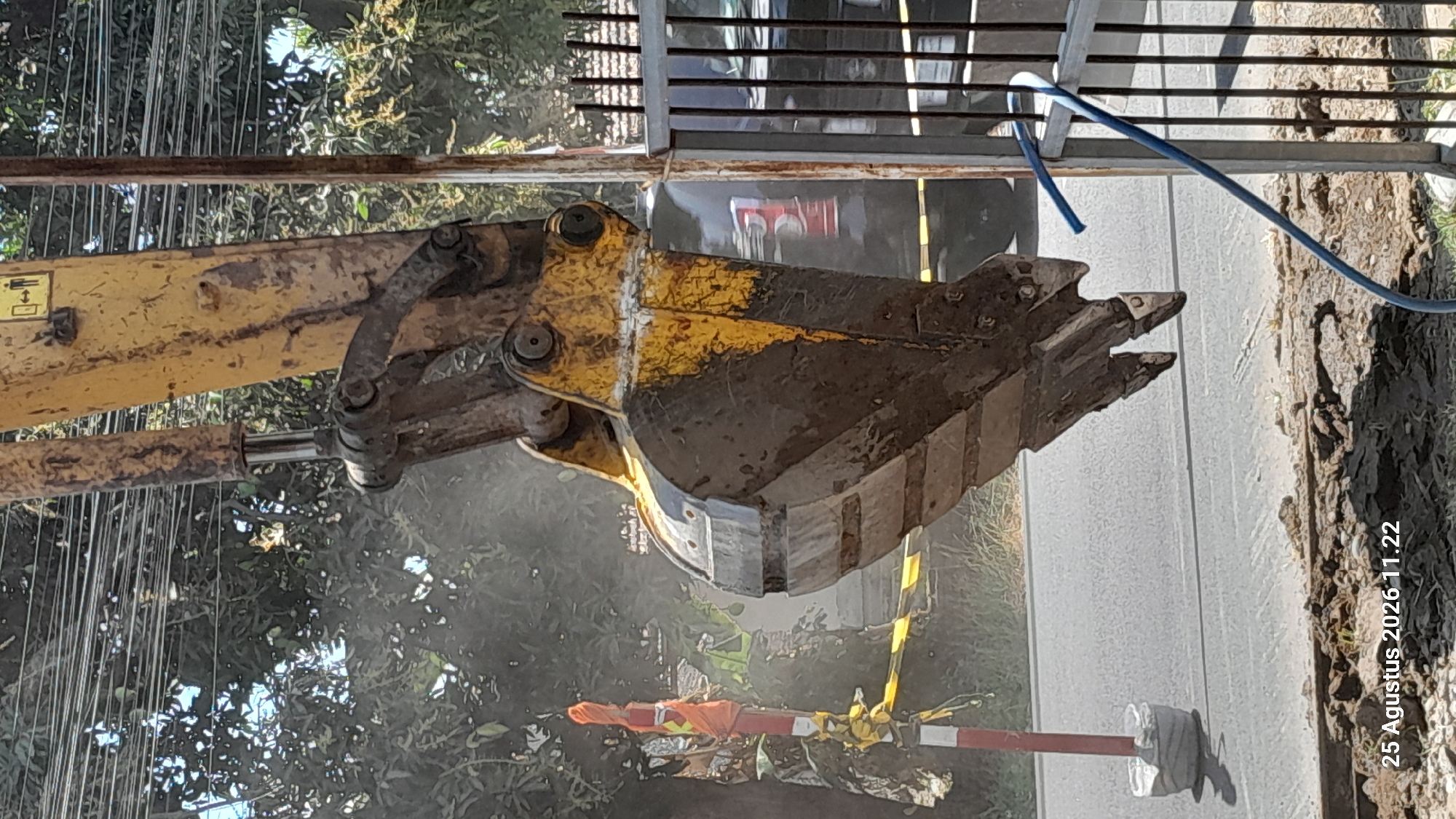 The image depicts an excavator bucket, heavily soiled with dirt, suspended in the foreground. The excavator is yellow with some faded paint. Behind the bucket, a hydraulic cylinder is visible, also exhibiting wear and dirt. The background shows a street with a striped traffic marker and yellow caution tape. Trees and power lines are present in the mid-ground, with a dark-colored vehicle partially visible behind a fence made of vertical bars on the right side of the image. A blue hose is draped over the fence. The timestamp "25 Agustus 2026 11.22" is present in the lower left corner, indicating it is a photograph taken on August 25, 2026, at 11:22 AM. The scene suggests ongoing construction or excavation work in an urban or suburban area, possibly in Jomblang, Indonesia, given the timestamp's date format and language.