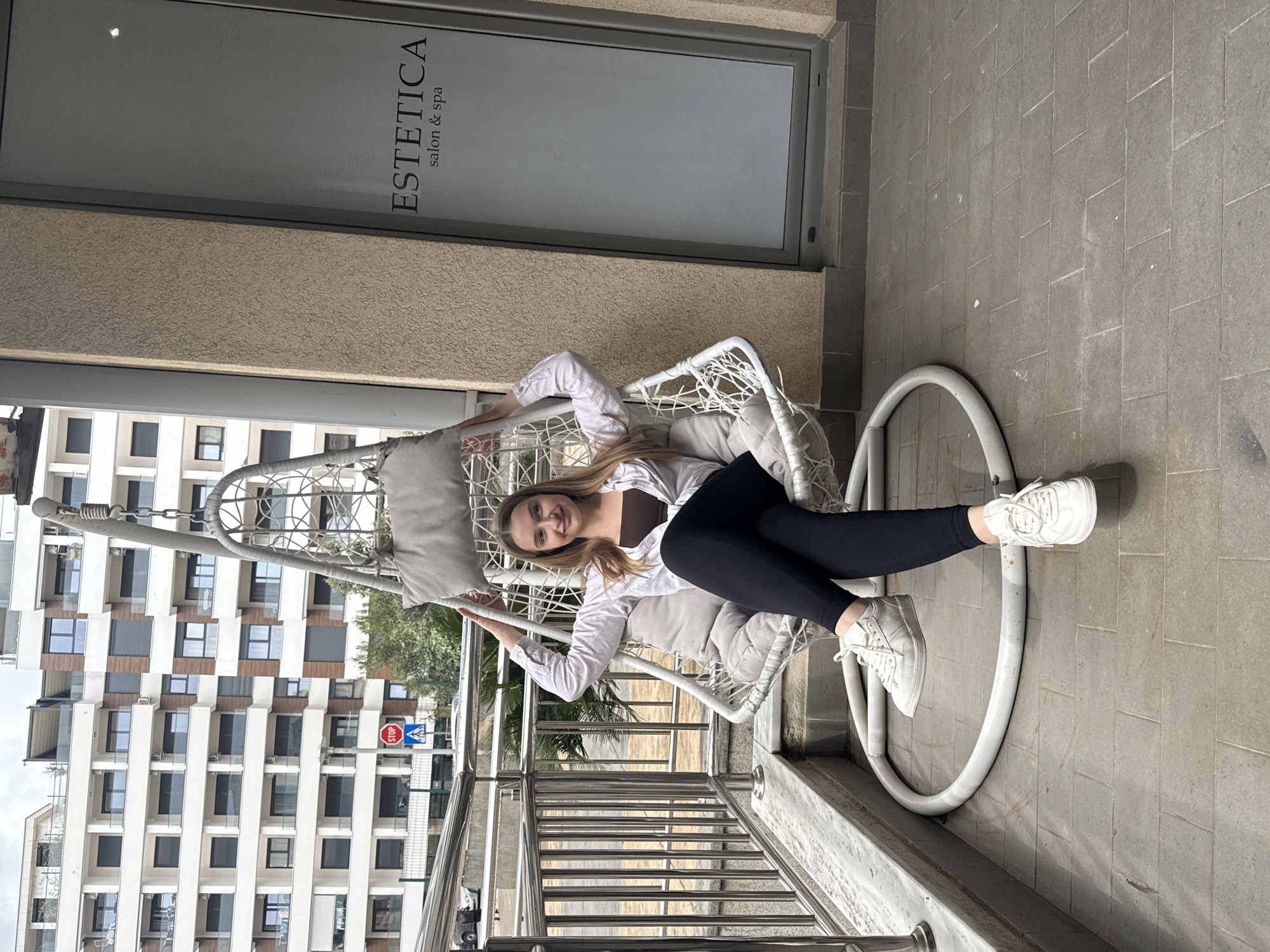 The image, rotated 90 degrees clockwise, depicts a young woman relaxing in a white, egg-shaped hanging chair on an outdoor terrace or balcony. She has long, light brown hair and is smiling brightly, looking directly at the camera. She is dressed in a white long-sleeved button-up shirt, a dark top underneath, black leggings, and white sneakers. The hanging chair features grey cushions for comfort.
In the mid-ground, a building with a textured light brown facade and a large, grey-framed window is visible. On this window, the text "ESTETICA" in large letters, with "salon & spa" below it, identifies the establishment. The floor of the terrace is made of grey tiles, and polished chrome railings are prominent in the foreground.
The background reveals a dense urban environment with multiple modern, multi-story buildings characterized by white facades and dark windows. Green trees provide a touch of nature amidst the city architecture. A red "STOP" sign and a blue "P" (parking) sign are discernible in the distance. The lighting suggests a soft, natural daylight, likely on an overcast or partly cloudy day, with no harsh shadows. The overall atmosphere is one of relaxation in an urban setting, consistent with the location context of Budva, Montenegro.