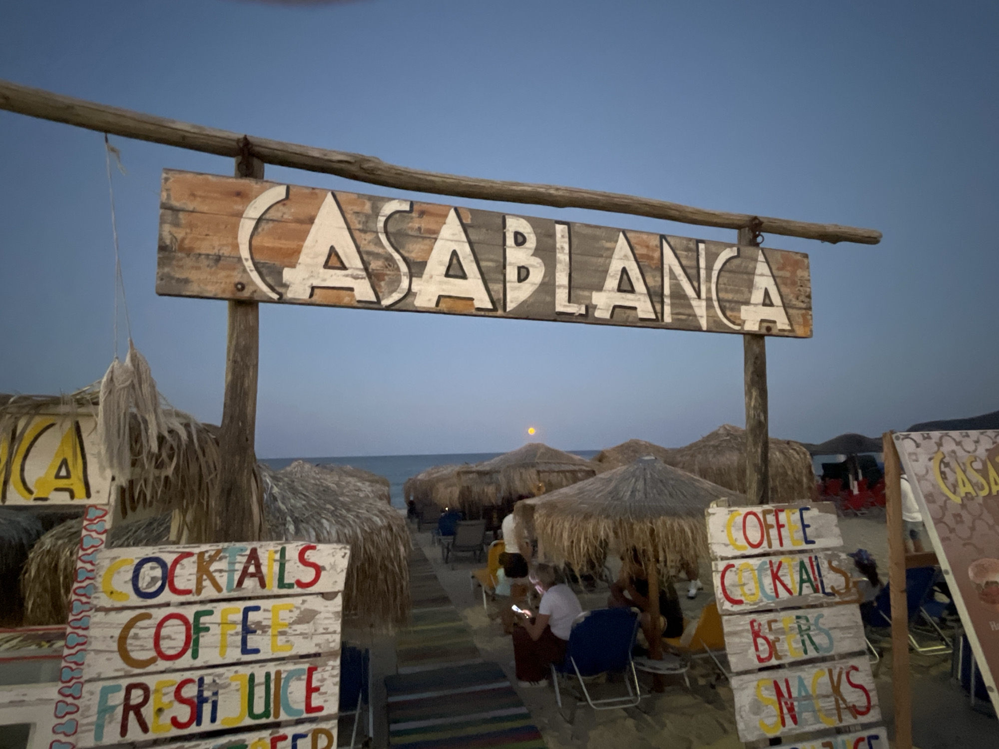The image depicts a beachside establishment in Toroni Municipal Unit, Greece, at dusk. A weathered wooden sign with "CASABLANCA" painted in white letters against a distressed wood background hangs from a rustic wooden frame, identifying the location. In the foreground, several smaller signs list offerings such as "COCKTAILS", "COFFEE", "FRESH JUICE", "BEERS", and "SNACKS", painted in colorful block letters on white, distressed wooden planks. Behind these signs, thatched umbrella-shaped roofs provide shade over seating areas on the sandy beach. A few people are seated under the umbrellas, some appearing to be patrons. The sea is visible in the background under a clear, pale blue sky, with a hint of a sunset or moonrise indicated by a faint orange glow on the horizon. The overall atmosphere suggests a relaxed, casual beach bar setting.