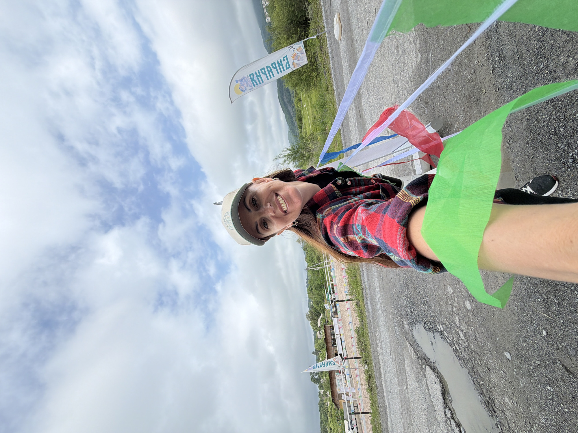 This is a selfie of a woman with long brown hair, wearing a red and black plaid shirt, a light-colored baseball cap, and a green streamer draped over her arm. She is smiling and looking directly at the camera. The photo is taken outdoors with a cloudy sky.
In the background, there is a road with puddles and cracks, suggesting it might have recently rained. There are also several flags and banners visible, some with colorful streamers, which could indicate an event or a race. One flag has text on it that appears to be in Cyrillic, reading "КЫЗЫЛ-ХАЙЯ". The surrounding landscape shows rolling green hills and trees.
The overall impression is of a cheerful moment captured during an outdoor activity, possibly a race or a festival, given the festive decorations and the woman's energetic pose. The weather appears overcast but not actively raining. The location context provided is Teploozersk, Russia.