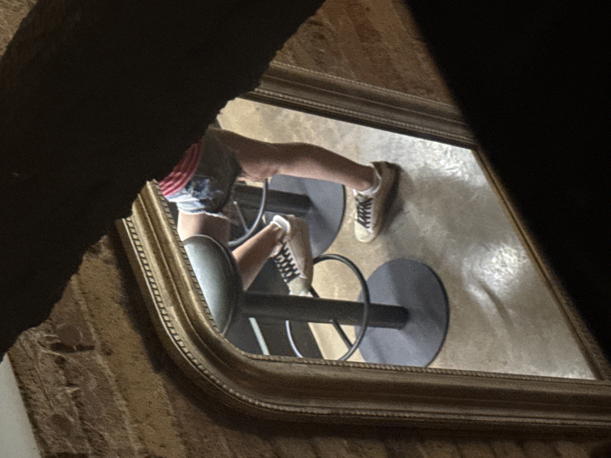 In Barcelona, Spain, a reflection in a gilded mirror shows a person seated on a black bar stool. The person is wearing denim shorts, a red and white striped top, and beige sneakers with black laces. The floor appears to be concrete. The mirror's frame is ornate and golden, positioned against a brick wall. The scene is dimly lit.