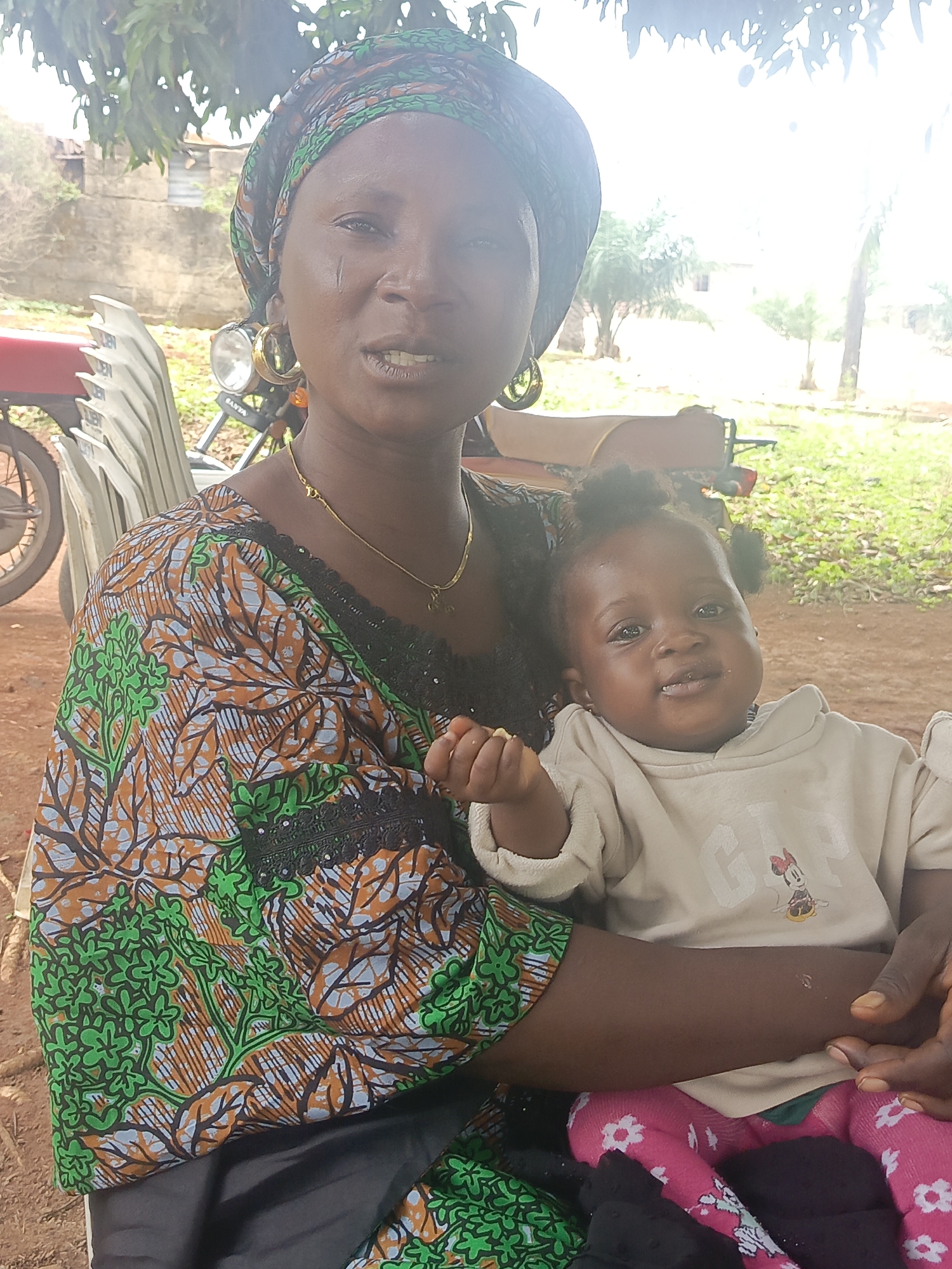 A mother and a baby are captured in a bright, outdoor setting in Andaha, Nigeria. The woman, with a dark complexion, is positioned centrally, holding the infant in her left arm. She wears a vibrant, patterned top featuring green, orange, blue, and black motifs with black lace trim, and a matching headscarf. Large gold hoop earrings and a thin gold necklace with a small pendant adorn her. Notable scarification marks are visible on her right cheek. Her mouth is slightly open, suggesting she might be speaking or smiling subtly.
The baby, also with a dark complexion, is seated comfortably in the mother's arm, looking directly at the camera with a gentle expression and small pigtails. The baby wears a light-colored, long-sleeved hooded top with the text "GAP" and a Minnie Mouse graphic visible on the front. Pink pants with white floral patterns cover the baby's legs. A small piece of food or an object is held in the baby's right hand.
The background reveals a casual outdoor environment with dirt ground, green foliage, and trees. Partial views of a red motorcycle with a visible headlight (text "SANTA") and white plastic chairs are seen behind the woman. A light-colored wall is present in the mid-ground. The scene is well-lit by natural daylight, indicating daytime.