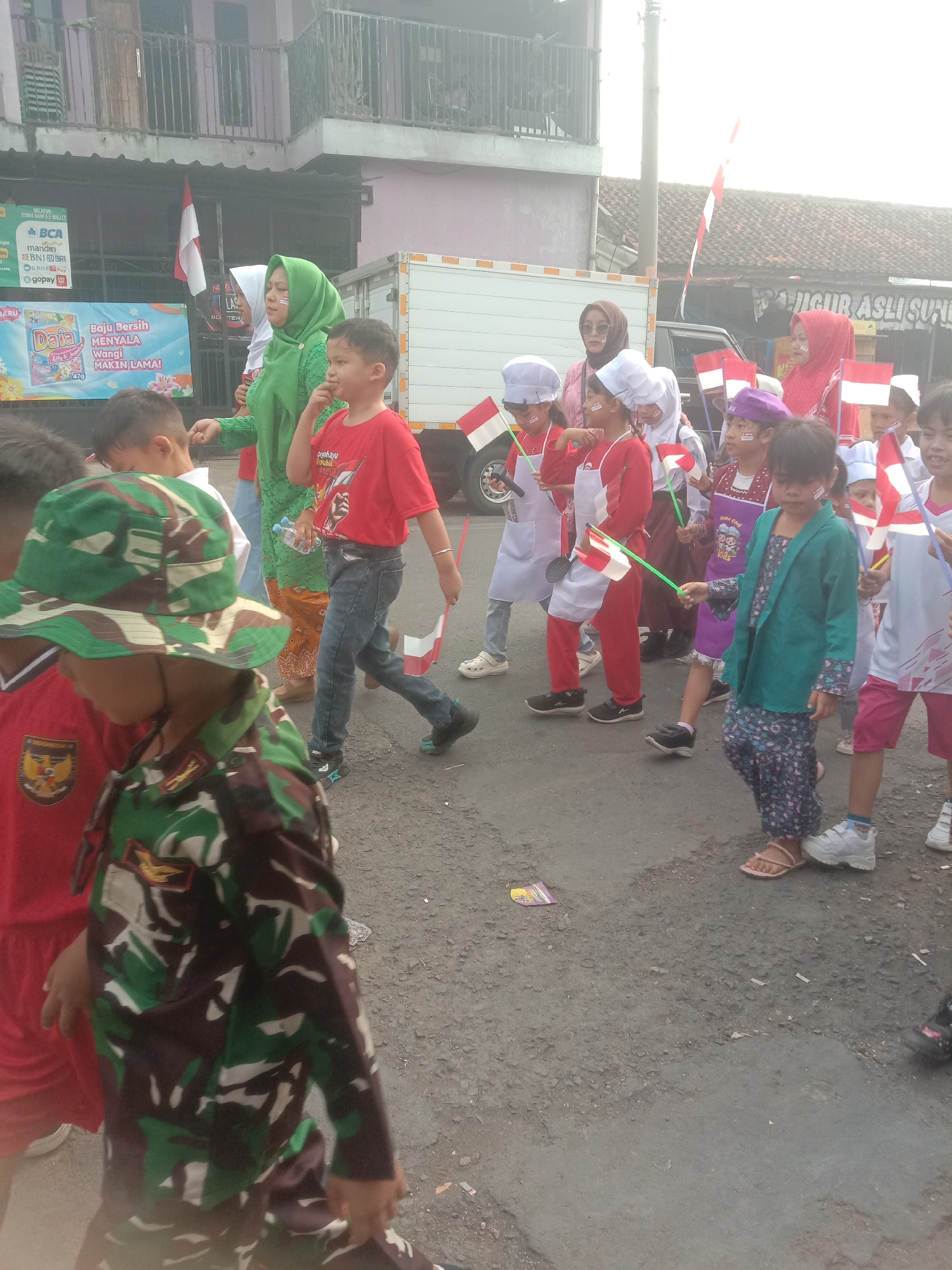 The image depicts a street scene in Bojong, Indonesia, likely during a parade or celebration, given the presence of children carrying Indonesian flags. The children are dressed in various outfits, including some in what appear to be chef hats and aprons, and others in camouflage military-style uniforms and hats. The adults are also dressed in traditional attire, with one woman wearing a green hijab and another in a red hijab. They are walking on a paved road with buildings on one side and a truck in the background. The overall atmosphere appears festive and communal.