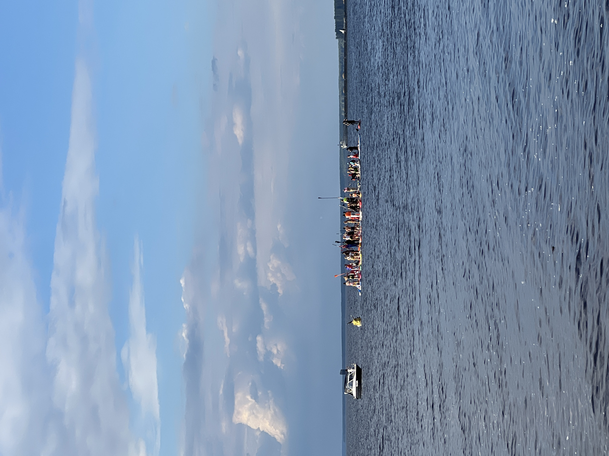 The image depicts a group of people participating in a paddleboard or kayak race on a wide body of water. The subjects are primarily human figures, each on their own paddleboard or in a kayak, forming a line that stretches across the frame. Some individuals are actively paddling, while others are positioned on their boards. There are also a few boats visible in the scene, including a small white motorboat in the foreground and a yellow inflatable object further back.
The setting is a large lake or river with calm, dark blue water that reflects the sunlight. The sky is a clear blue with scattered white clouds, suggesting a bright and possibly warm day. The time of day appears to be late morning or early afternoon. The location is identified as Chaikovsky, Russia. There are no discernible emotions visible from the subjects due to the distance and the orientation of the image. No visible text is present in the image. The overall activity is clearly a water sport event, likely a race or a group paddle.