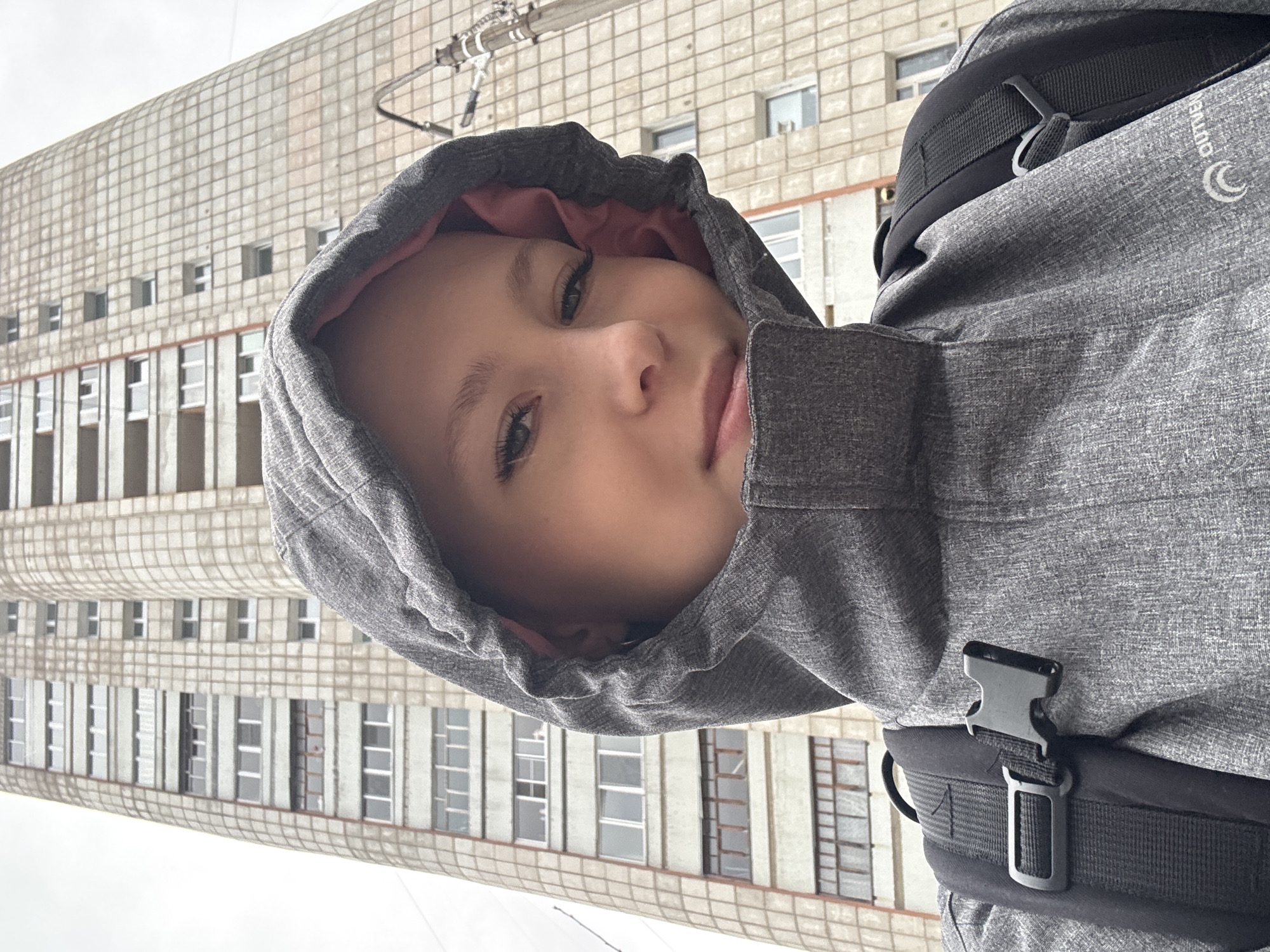 A child with light skin, wearing a gray hooded jacket with a pink lining, looks into the camera. The jacket is part of a backpack, visible by the straps and buckles. The background is an exterior view of a multi-story building made of light-colored concrete, with numerous rectangular windows and vertical columns. The scene appears to be in Chaikovsky, Russia, based on architectural and environmental context.