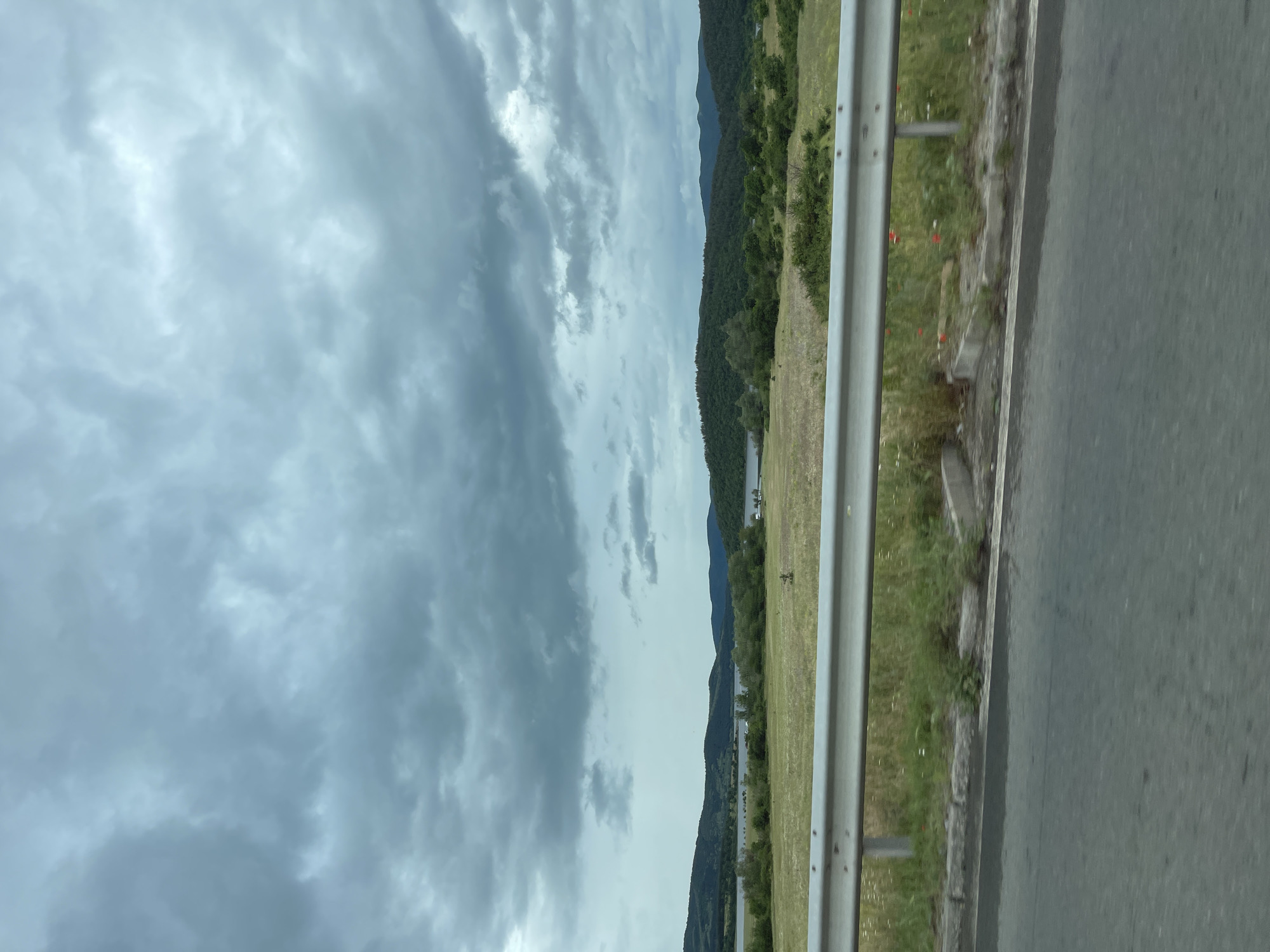 A horizontal outdoor shot taken from a moving vehicle shows a cloudy sky, a landscape with trees and grass, and a gray asphalt road. A metal guardrail runs along the side of the road. In the distance, there is a body of water. The location is identified as near Konare, Bulgaria.