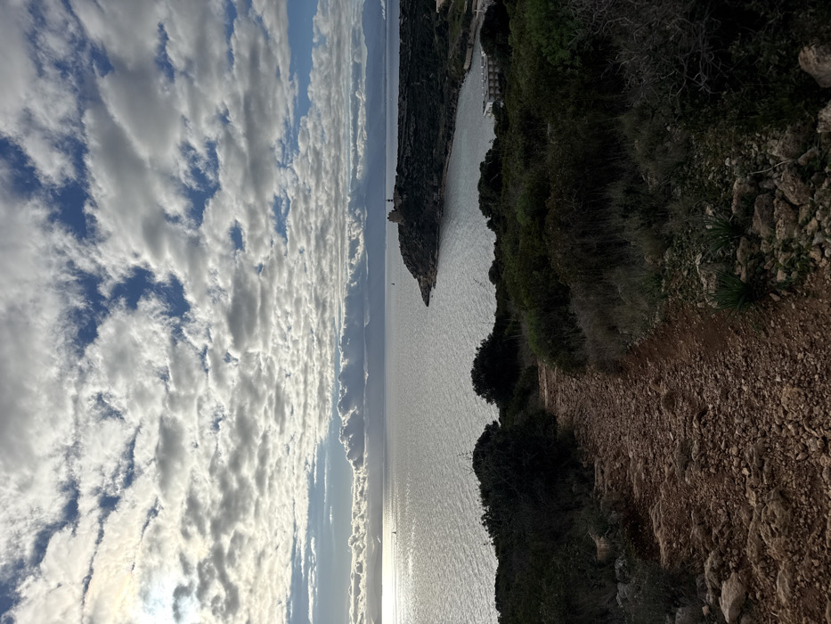 The image displays a coastal landscape captured from an elevated viewpoint, specifically identifying as the Capo Sant'Elia promontory in Cagliari, Italy, looking eastward over the Gulf of Cagliari.
The foreground is dominated by a reddish-brown dirt path composed of loose soil and small rocks, leading downwards and slightly curving right. Sparse, dry scrub vegetation and small bushes flank this path.
The midground features a shimmering expanse of the Mediterranean Sea under a partly cloudy sky. The water reflects the bright light from the left side of the frame, where the sun is partially obscured but brightly illuminates a section of the sky and water. Two small sailboats are visible on the water: one in the distant left-center and another smaller one closer to the right. A prominent vegetated headland, Capo Sant'Elia, extends into the sea from the right, forming a bay. On the highest point of this headland, an ancient-looking stone tower structure, likely the Torre di Sant'Elia, is visible. Below the tower, within the bay and partially obscured by vegetation, a light-colored, multi-story building with a flat roof, possibly a hotel or residential complex, is situated near the waterline.
The background shows faint outlines of distant landmasses or mountains across the gulf under a horizon line. The sky above is characterized by an extensive pattern of cumulus and stratocumulus clouds, with patches of clear blue sky visible between them. No individuals are present in the scene.