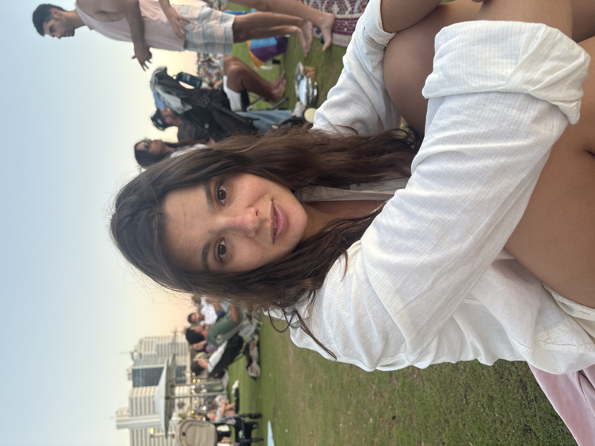 A young woman with long, dark hair and freckles is seated on the grass, facing the camera. She is wearing a loose-fitting white linen shirt. Her arms are crossed in front of her, and her legs are bent. In the background, several other people are visible, some sitting on the grass or in chairs, enjoying a picnic. The setting appears to be a park or outdoor gathering area. In the far background, tall buildings are visible, suggesting an urban environment. The sky is clear and light, indicating it is likely late afternoon or early evening. The overall atmosphere is relaxed and casual. The location appears to be Tel Aviv, Israel, given the characteristic architecture visible in the background.