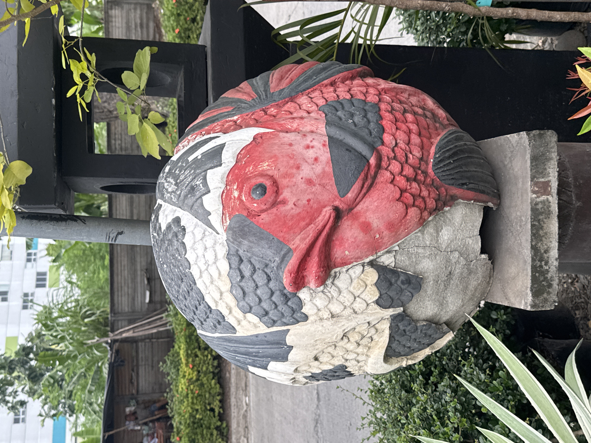 The image shows a weathered, spherical sculpture of a fish, predominantly red with black and white patterned scales, placed on a concrete pedestal. The sculpture is positioned outdoors, with a blurry background that includes greenery, a wooden fence, a building with white and teal stripes, and a paved area resembling a road or sidewalk. A dark, geometric structure with an opening is visible behind the sculpture. The lighting suggests it is daytime.