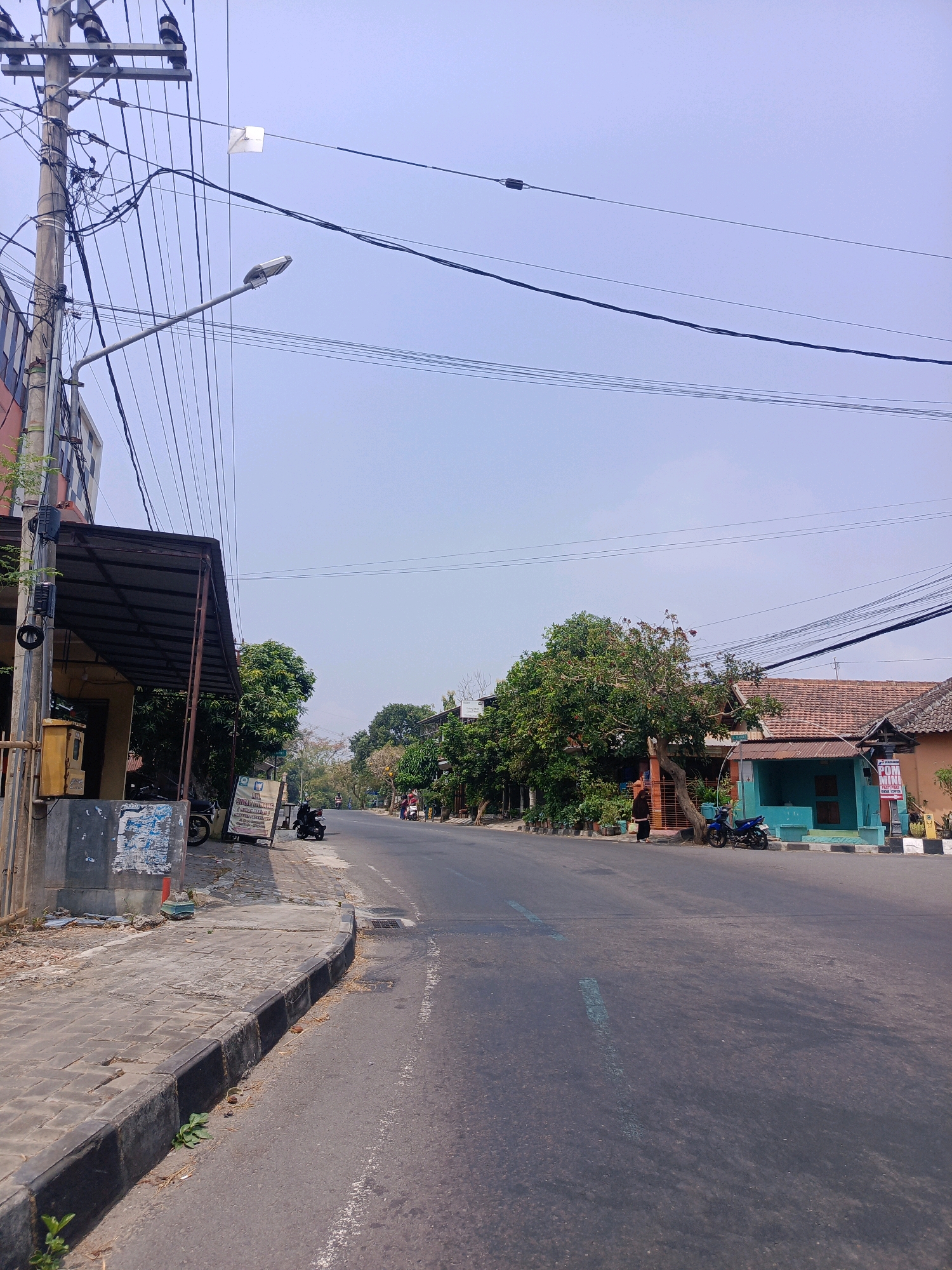 The image shows a street scene in Mangkujayan, Indonesia, under a pale blue sky with a haze. A paved road runs diagonally from the lower right to the upper left of the frame. To the left of the road is a raised sidewalk with a dark curb. Alongside the sidewalk, there is a building with a metal awning, electrical poles with numerous wires, and a yellow mailbox. Further down the sidewalk, a sign displays text in Indonesian. Beyond the sidewalk and road, there are trees and several houses with tiled roofs. Two motorcycles are parked on the side of the road. In the distance, a few people are walking or riding motorcycles.