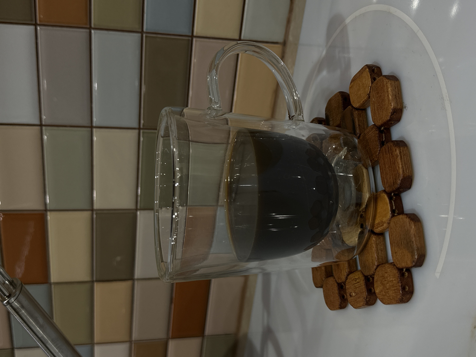A close-up shot shows a glass mug filled with a dark liquid, possibly coffee or tea, sitting on a wooden coaster composed of small, connected wooden pieces. The mug is a double-walled design, providing insulation. The coaster is positioned on a white surface, likely a countertop or table. The background features a tiled wall in a variety of neutral colors, predominantly beige, gray, and brown. The scene appears to be indoors, possibly in a kitchen or cafe setting. The location in Stara Zagora, Bulgaria cannot be determined from the image.