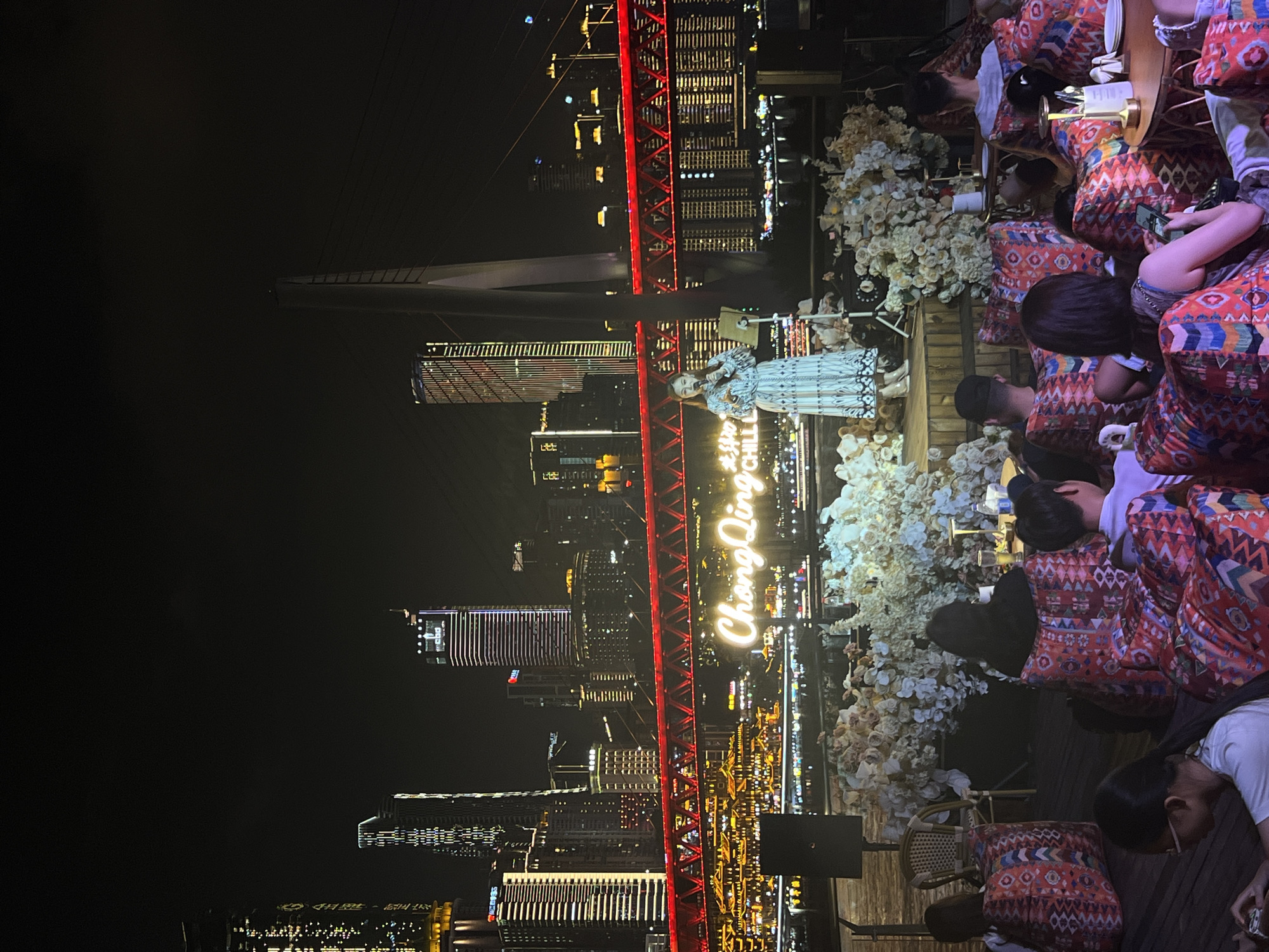 A wide shot captures a lively evening scene at a riverside venue, likely in Chongqing, China, possibly near Jiangbeicheng Street, given the distinctive cityscape. In the foreground, a group of people, diverse in age, are seated on colorful, patterned beanbag chairs, facing a raised stage. Their attention is directed towards the stage, where a woman in a long, patterned dress is singing into a microphone. She stands beside a music stand and a speaker, bathed in the warm glow of stage lights and surrounded by abundant white and cream floral arrangements. Behind the stage, a prominent neon sign, illuminated in a bright white, reads "Chongqing Chill." Further in the background, a vibrant red bridge spans the river, its intricate metal structure illuminated, contrasting with the dark night sky. The bridge is a striking element, and its reflection shimmers on the water below. Beyond the bridge, the iconic Chongqing skyline is visible, a dense cluster of skyscrapers adorned with countless twinkling lights, creating a dazzling urban panorama. The overall atmosphere suggests a popular entertainment spot, likely a bar or restaurant, offering live music and city views.