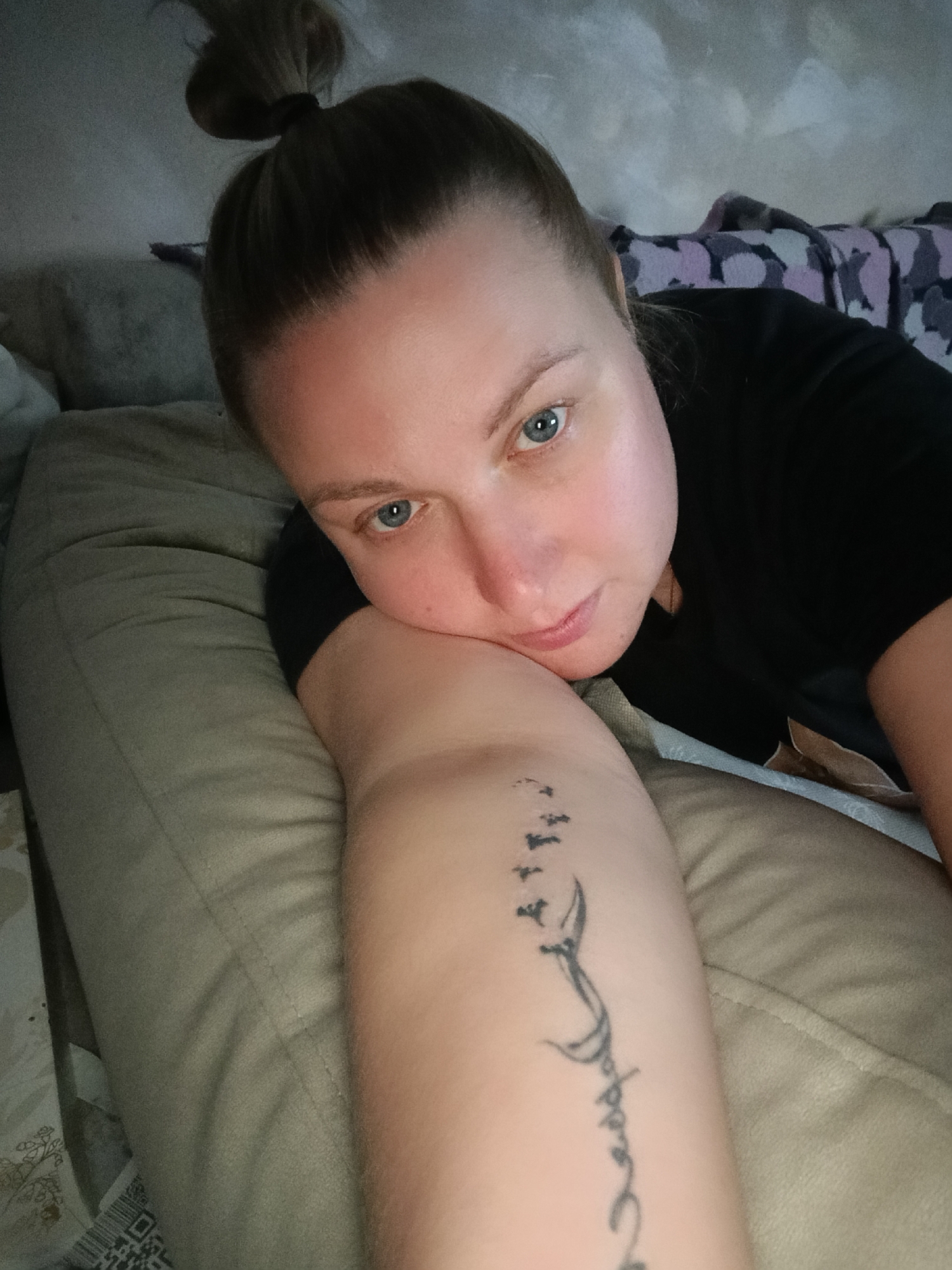 A medium, eye-level shot depicts a light-skinned woman with blue eyes, her hair pulled back in a bun, lying on a bed or sofa. She is wearing a black top. Her right arm is positioned in the foreground, supporting her head as she looks directly at the camera. A tattoo of birds in flight above a script is visible on her inner forearm. The scene appears to be indoors, in what looks like a private residence in Chaikovsky, Russia. Pillows and a patterned blanket are visible in the background.