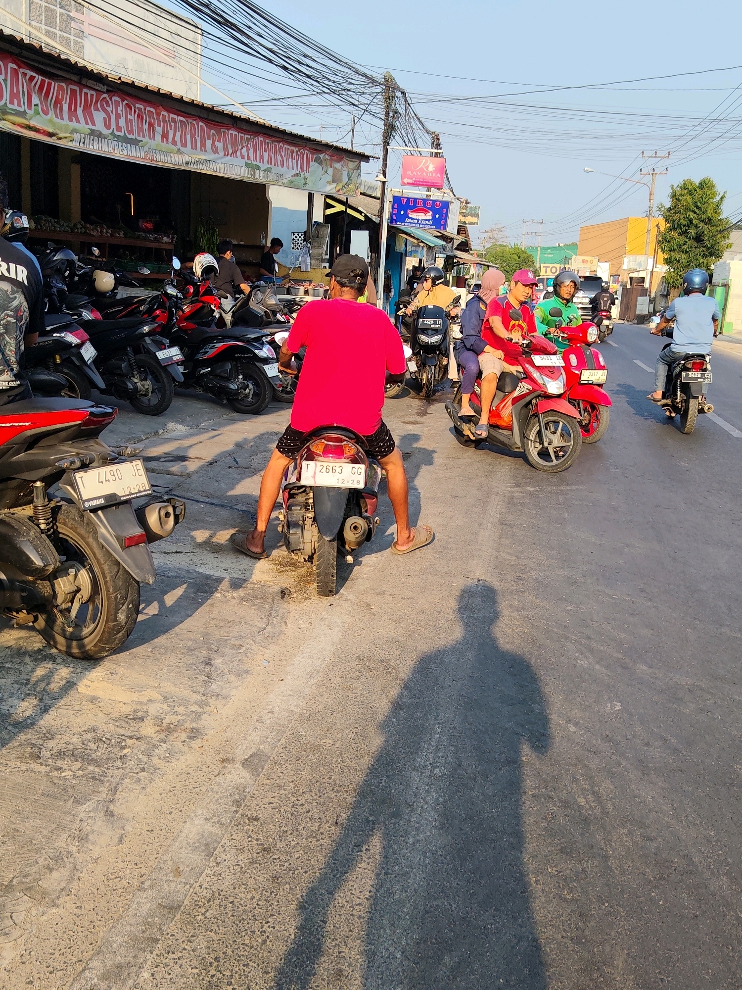 The image captures a street scene in Purwakarta, Indonesia, during daylight hours. In the foreground, the back of a person wearing a bright red shirt and dark shorts is visible, seated on a scooter. The scooter has a license plate reading "T 2663 GG". To the left of this scooter is another scooter with the license plate "T 4490 JE". A variety of other motorbikes are parked or being ridden in the background. A building with a large banner displaying "SAYURAK SEGAR AZURA" and "MENERIMA PESANAN" is situated on the left side of the image, suggesting a market or food stall. Further down the street, more buildings, signs, and people on scooters are present, indicative of a bustling urban environment. The sky is clear, and the sun casts distinct shadows on the asphalt road. Overhead, numerous electrical wires are strung across the scene.