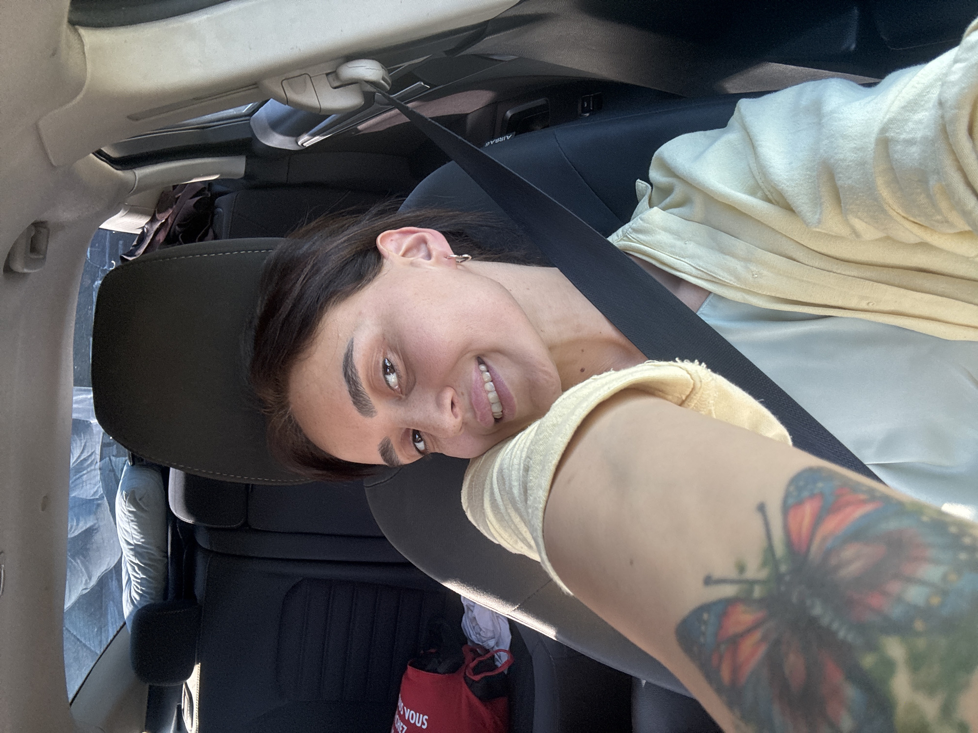 The media file is a selfie taken by a young woman inside a car. She is leaning back against the headrest, looking at the camera with a smile. She has dark hair, brown eyes, and is wearing a light yellow top. A seatbelt is visible across her chest. Her left arm, which is extended towards the camera, has a tattoo of a butterfly on it.
The setting appears to be daytime, as evidenced by the natural light coming from the car's sunroof. The interior of the car is visible, including the seats and the car's roof lining. A red bag with white text is visible in the back seat.
The image conveys a casual and relaxed mood. The woman's smile and direct gaze suggest a happy or playful moment. The presence of the car indicates she is likely traveling. The location context of Tbilisi, Georgia, is provided externally and is not directly discernible from the visual elements of the image itself.