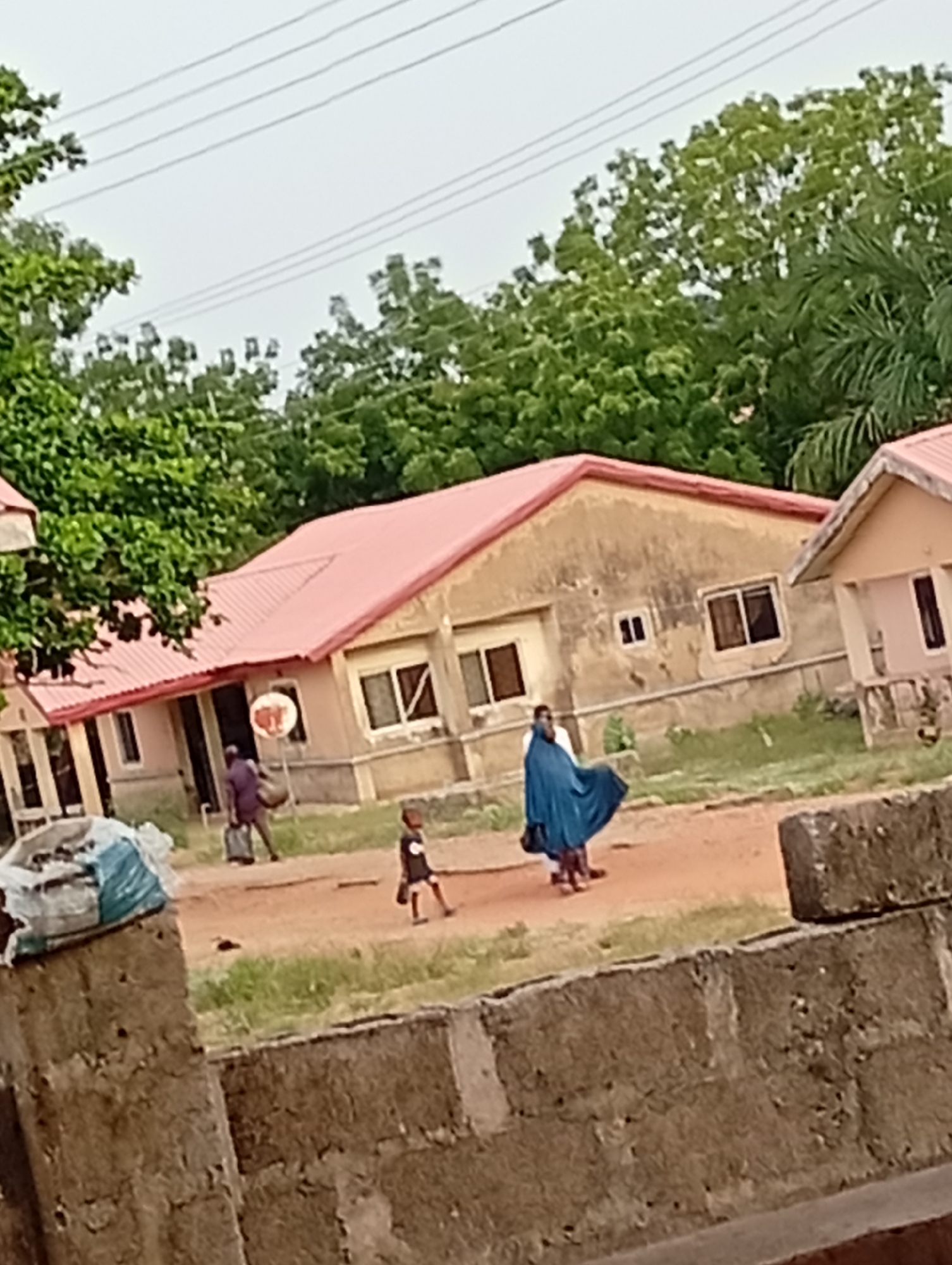 A street scene shows a few people walking on a dirt road in front of houses. There is a woman in a blue dress and a child to the right. Another person carries a bag to the left of the image. The sky is overcast. The location is unknown.
