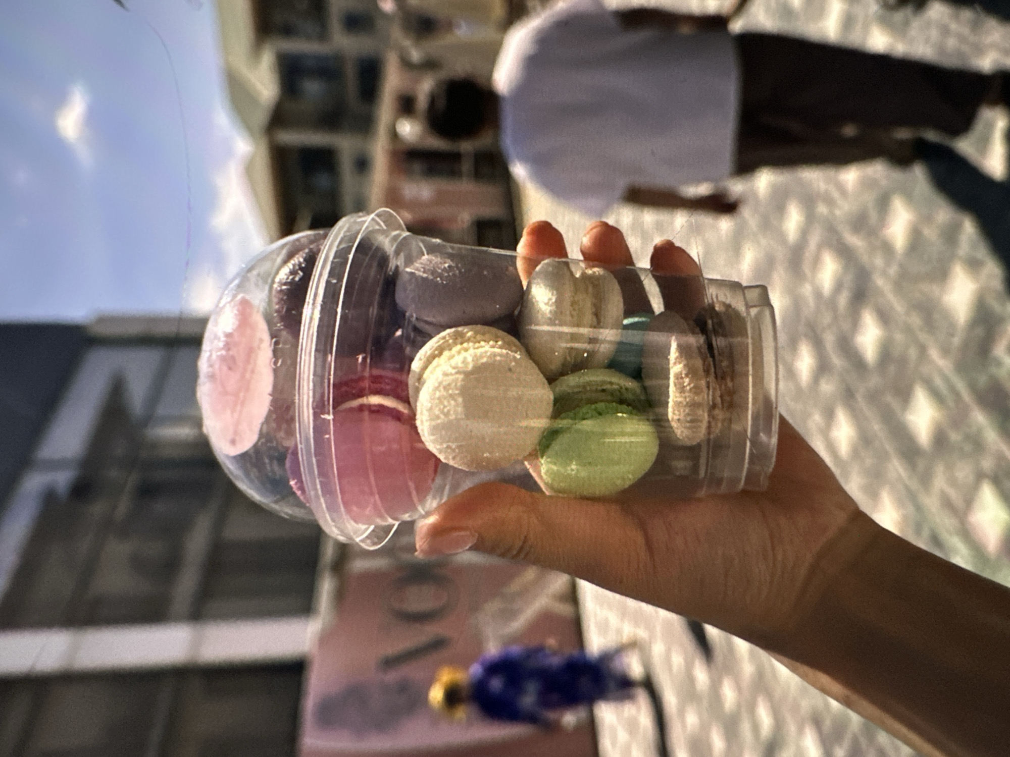 In an outdoor urban environment, likely within the city of Кызыл-Булак, Kyrgyzstan, a person's hand holds a clear plastic cup filled with an assortment of macarons. The macarons display a variety of pastel colors including pink, purple, cream, and green. Behind the cup, the background suggests a street scene. To the left, a person in a blue robe and a yellow hat is visible, walking away from the viewer, with a large sign that appears to read "VOGUE" in the mid-ground. To the right, another person, seen from behind, is wearing a white t-shirt and dark pants, walking away. The ground is paved with a pattern of lighter and darker square tiles. The sky above is blue with scattered white clouds, and the lighting indicates it is daytime, likely sunny.