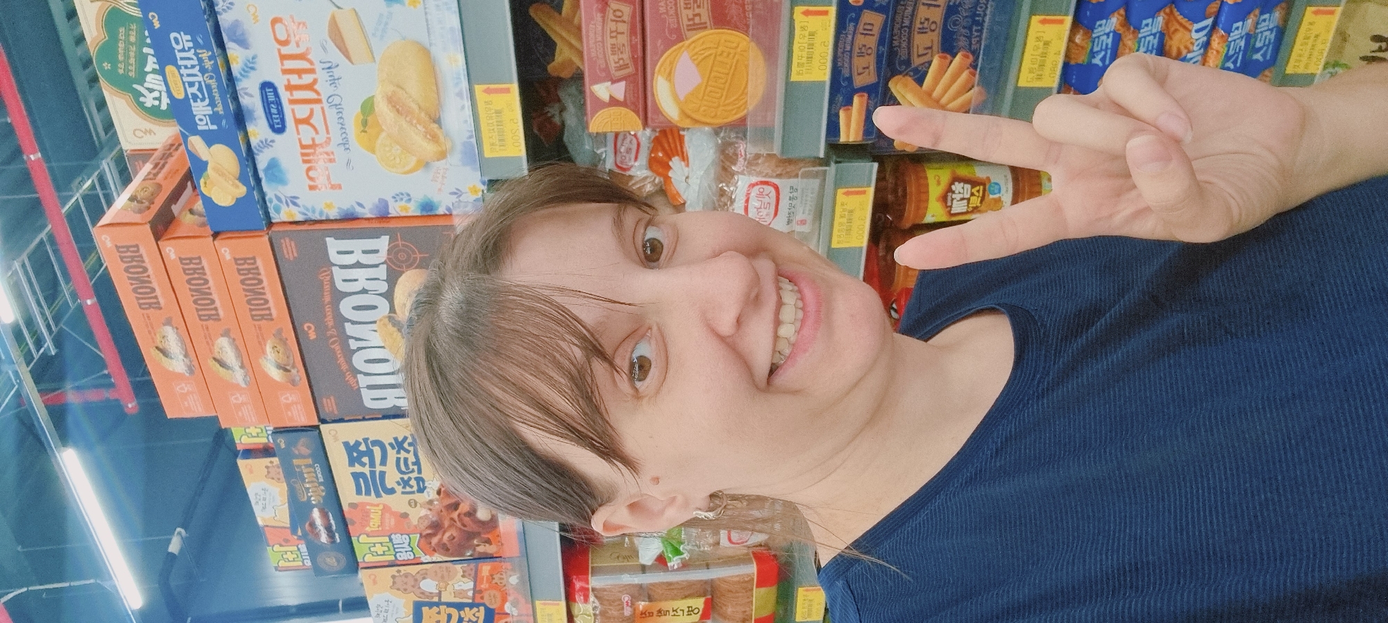 A person with light skin, brown hair with bangs, and wearing a dark blue shirt is taking a selfie with a peace sign. They are positioned in front of a shelf stocked with boxes of snacks, cookies, and confectionery goods, many with Korean labeling. The location is suggested to be within Dunnae-myeon, South Korea.