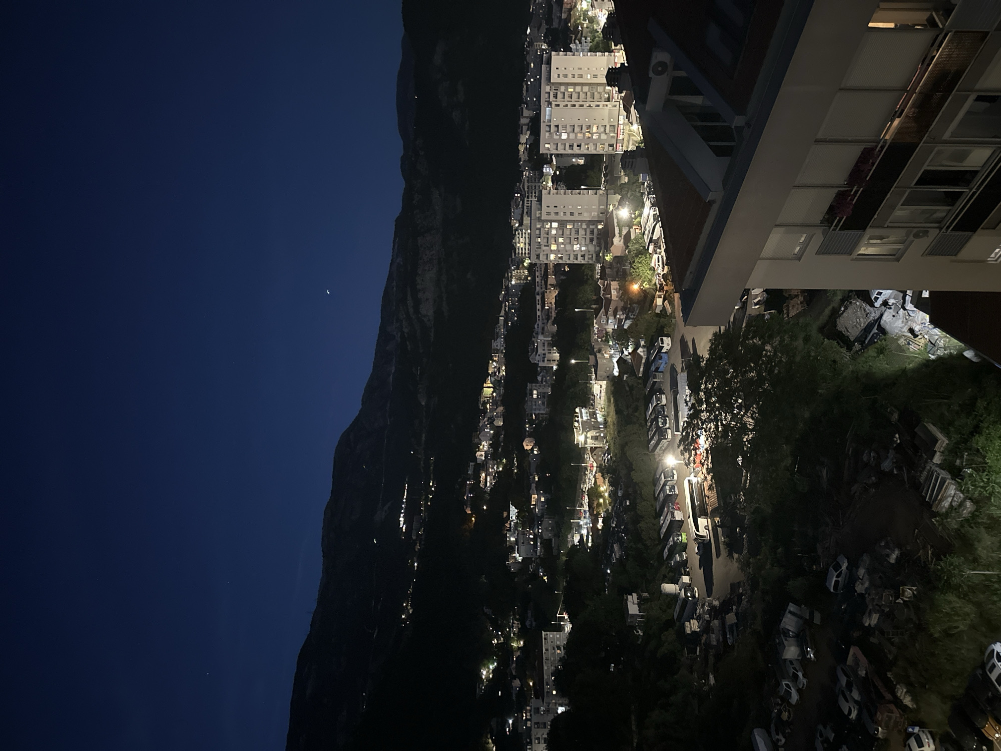 A nighttime aerial view captures a segment of Budva, Montenegro, revealing a landscape dotted with urban development and natural features. The sky, a deep navy blue, is sparsely populated with a few faint stars and a sliver of the moon. Dominating the background is a dark, mountainous silhouette, with scattered lights indicating settlements or structures along its slopes.
In the midground, a collection of buildings, primarily modern apartment blocks with illuminated windows, forms a part of the cityscape. These structures vary in height and architectural style. Below them, a busy street or parking area is visible, illuminated by streetlights, with numerous vehicles, including buses and cars, parked or in motion. Some of these vehicles appear to be parked in a depot or bus station.
The foreground is occupied by the edge of a building, partially obscuring the view. Below this building, an area with visible debris, possibly construction materials or discarded items, is present, surrounded by trees and vegetation. The overall impression is a lively urban area transitioning into a mountainous backdrop under the cloak of night.