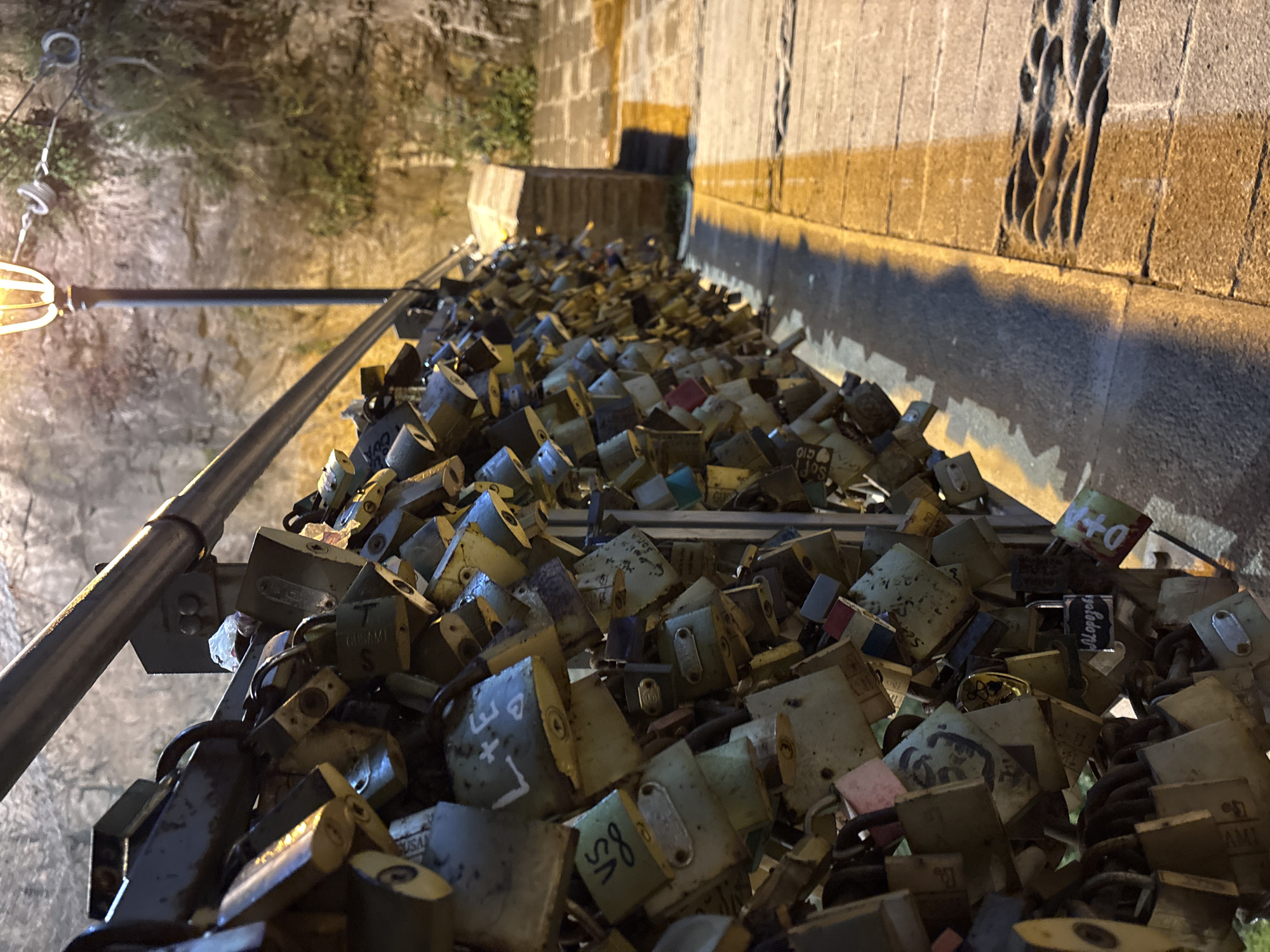 A close-up, eye-level shot captures a railing densely covered with padlocks. The padlocks are of various sizes and colors, with some appearing newer and others showing signs of wear and tear. Many of the padlocks have inscriptions or drawings on them, suggesting they are love locks. The railing itself is metallic and appears to be part of a bridge or walkway. In the background, a lamppost with a glowing bulb is visible, casting a warm light. Behind the lamppost, a textured stone wall or cliff face is illuminated, with some greenery growing on it. The ground in the foreground is paved with stone, and a strip of golden light from the lamppost cuts across it, creating a shadow from the railing and padlocks. The overall scene suggests a romantic or sentimental location, likely a public space where couples attach locks as symbols of their commitment.