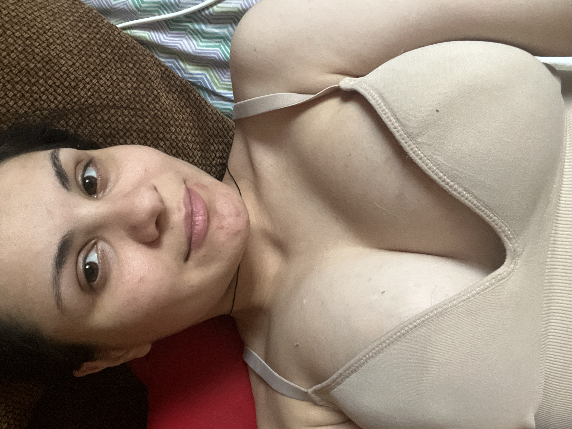 The media file shows a close-up, overhead shot of a person lying down. The person's face is partially visible on the left side of the frame, with their eyes open and looking towards the camera. They have dark eyebrows and lips. The person is wearing a beige bra. The background consists of a brown textured surface and a striped fabric in shades of green, blue, and white. The lighting appears to be artificial, suggesting an indoor setting. There are no visible signs of text in the image. The overall impression is a casual, personal photograph.