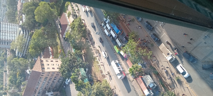 This high-angle, aerial photograph depicts a busy urban streetscape in Nairobi, Kenya, specifically focusing on University Way, observed from an elevated viewpoint within a building. The scene is bathed in bright daylight, casting defined shadows.
The central feature is a multi-lane asphalt road with significant vehicular traffic flowing in both directions. The lanes closer to the viewer show moving vehicles, including private cars (dark-colored sedans, SUVs, and a white car), a white flatbed truck, and a large reddish-brown bus. The mid-ground lanes exhibit substantial congestion, with numerous public service vans (matatus) of various colors (white, blue, green, multi-colored) queued alongside private cars and a white bus with red accents. Motorcycles are also intermittently visible between vehicles.
Pedestrians are present along the sidewalks bordering the road, particularly on the right side, with individuals walking and some appearing to cross. A low barrier or fence separates different sections of the road.
To the immediate left, a large, multi-story building, likely part of the University of Nairobi's Main Campus, features a light brown/beige facade, a prominent reddish-brown hip roof, and numerous small, rectangular windows. Behind this, and across the street, smaller buildings with similar reddish roofs are interspersed with dense green foliage. In the background, a tall, modern grey high-rise building stands, along with another building exhibiting a white and dark striped facade.
Abundant green trees and vegetation are prevalent throughout the scene, lining roads, occupying green spaces, and surrounding buildings. A fenced green area is visible to the left of the main road. A partial window frame is visible along the far right edge of the image. The distant sky appears slightly hazy.