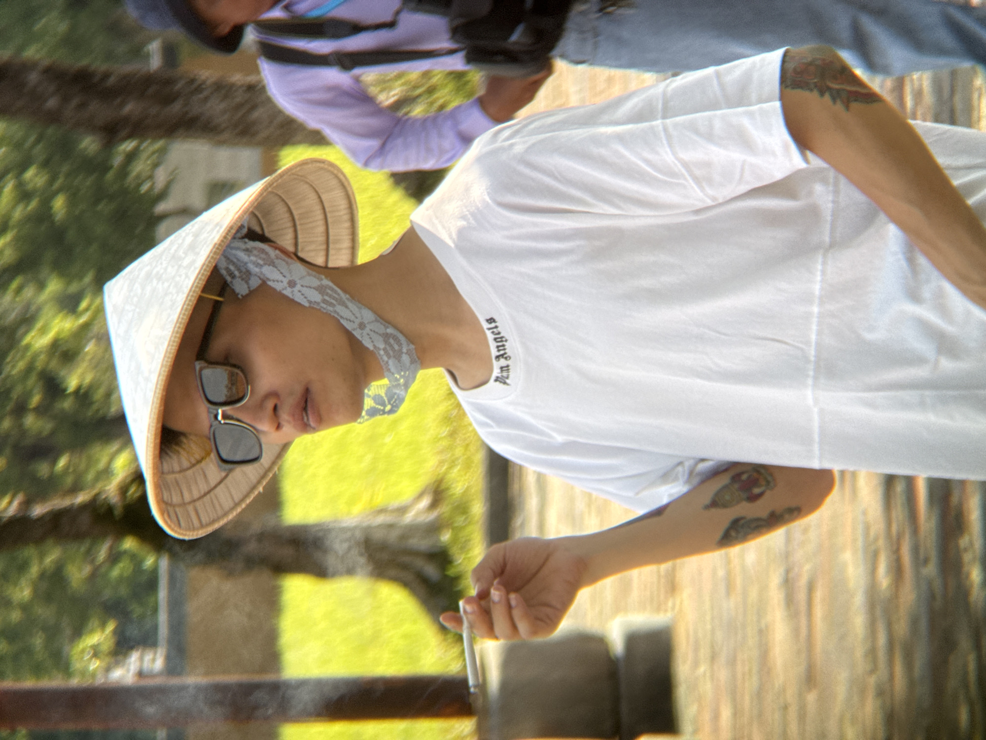 A medium shot shows a person wearing a conical hat and sunglasses, smoking a cigarette. The person is also wearing a white t-shirt with "Palm Angels" printed on the collar. The person has tattoos on their arms. In the background, a person in a purple shirt and a hat is visible. The scene appears to be outdoors with trees and a paved walkway. The location is identified as Phú Xuân Ward, Vietnam.