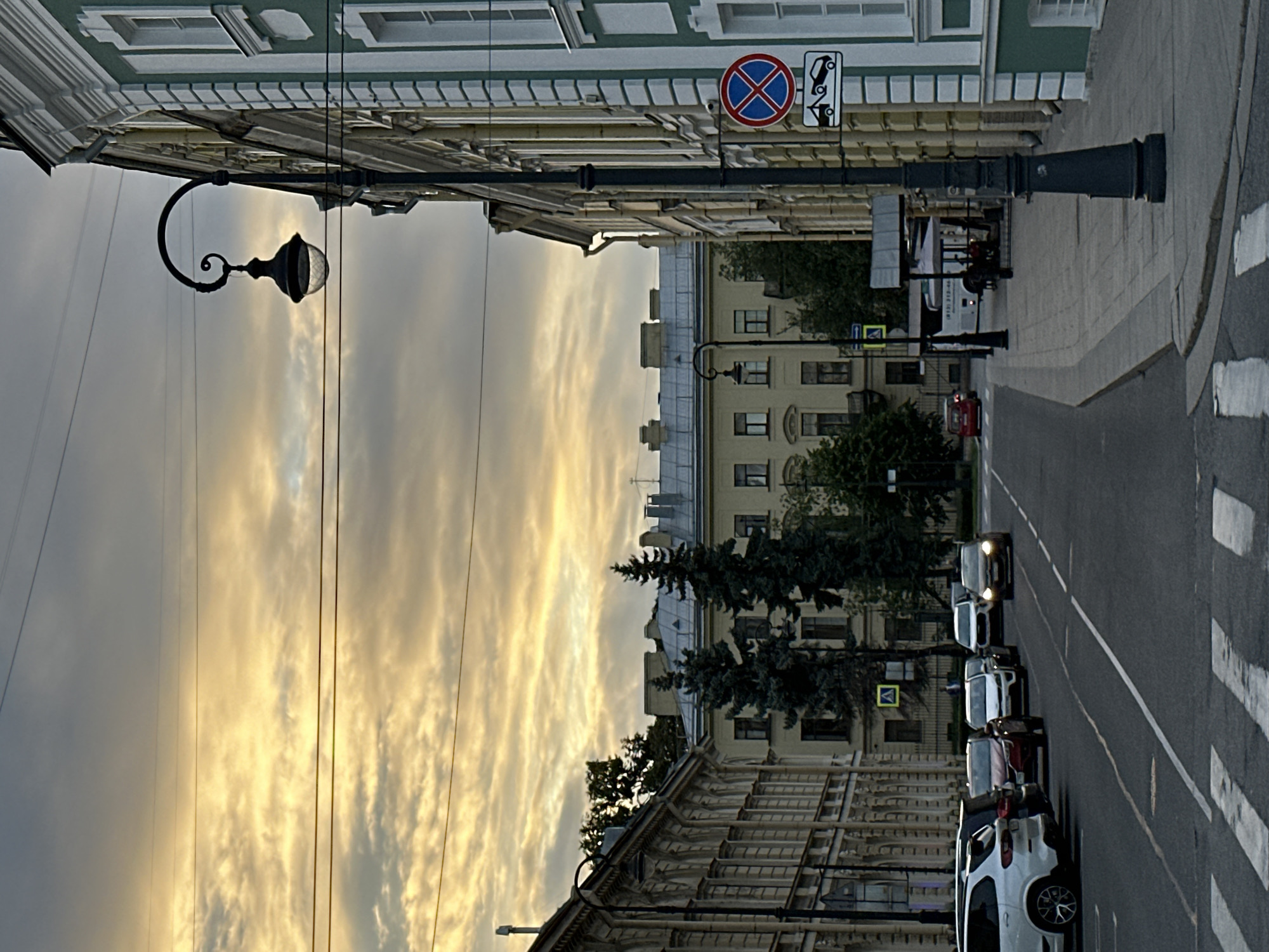 The image depicts a street scene in Saint Petersburg, Russia, likely during the daytime given the bright sky. The sky is filled with dramatic, golden-hued clouds, suggesting either sunrise or sunset.
On the right side of the frame, a tall, dark lamppost with an ornate curved arm stands prominently. It is positioned next to a building with a light green facade and white decorative molding. A traffic sign is attached to the lamppost, displaying a red circle with a blue diagonal cross and a tow truck symbol below it, indicating no parking or stopping and that vehicles will be towed.
To the left of the street, a long building with ornate architectural details, possibly stucco facades and arched windows, lines the sidewalk. Further down the street, other buildings are visible, including a pale yellow building with multiple windows and a grey tiled roof. Two tall, dark green evergreen trees stand in front of this yellow building.
The street itself is paved and appears to be a multi-lane road. Several cars are parked on the left side and driving on the right side of the road. White crosswalk lines are visible at the bottom of the image. A white bus with some text and numbers on its side is parked or moving slowly on the right side of the street, near a covered outdoor seating area. The overall atmosphere is urban, with a mix of historical architecture and modern elements.