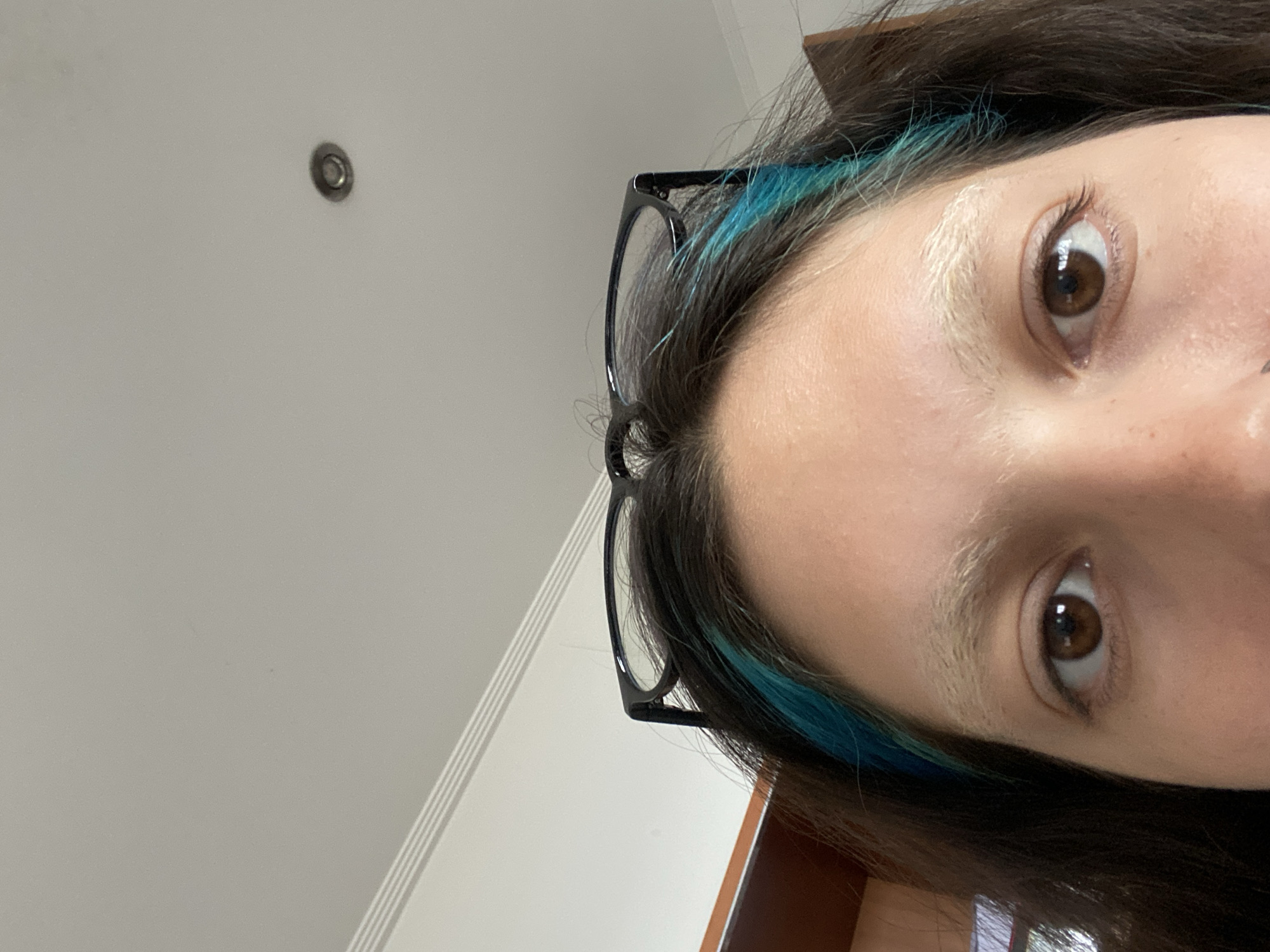 The image is a close-up, tilted selfie of a person's face. The person has brown eyes, fair skin, and dark brown hair with streaks of blue. They are wearing black-rimmed glasses. The background is a light-colored wall with a ceiling light fixture and part of a wooden door. It is not possible to ascertain if the location is Gijón/Xixón, Spain, as the scene provides no location-specific clues.