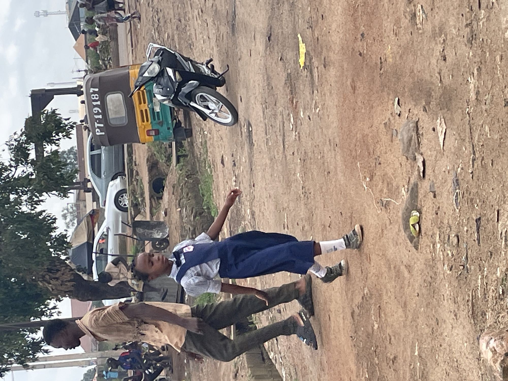 The image captures a scene in Bukuru, Nigeria, with a focus on two individuals. In the foreground, a child wearing a blue school uniform with a white collared shirt underneath is seen in motion, with their right arm outstretched. They are wearing white socks and dark sneakers. Beside the child, an adult in a light brown shirt and dark trousers is walking.
In the background, a motorized tricycle, likely a "Keke Napep" for local transport, is visible. It is a multi-colored vehicle with a registration plate reading "PT-19187". A motorcycle is parked next to the tricycle. Further in the background, other people are scattered around, and a white car is parked. Trees and some structures, possibly buildings or market stalls, complete the setting.
The ground is a dirt or gravel surface, common in many areas of Nigeria. The lighting suggests it is daytime, possibly mid-morning or afternoon, with diffused light indicating an overcast sky or a hazy atmosphere. The scene appears to be an outdoor, everyday environment, possibly a street or a market area. There are no explicit cues about the emotions of the individuals, but the child's pose suggests movement or play. The overall impression is a snapshot of daily life in a Nigerian town.