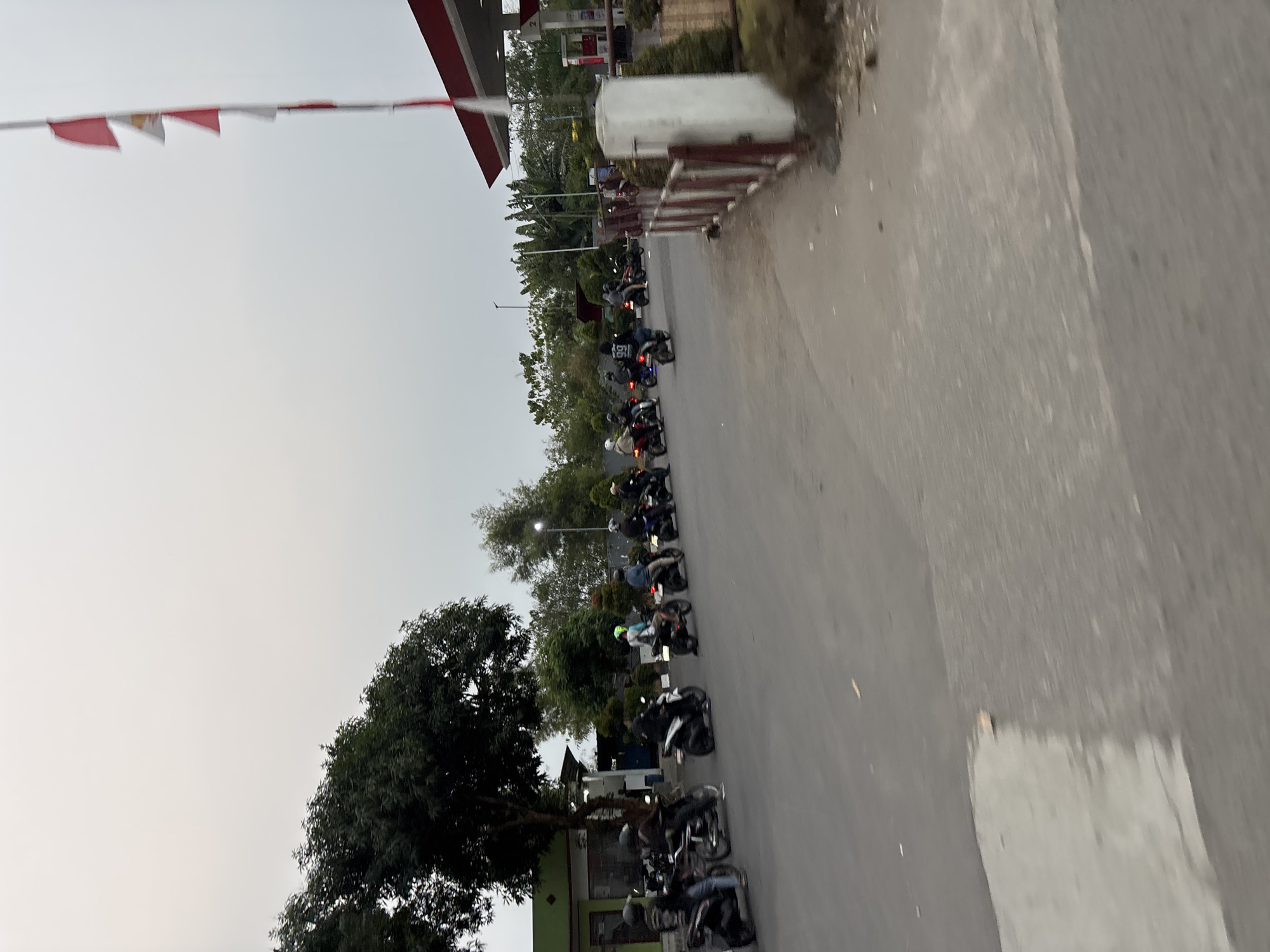 The image depicts a gas station in Bekasi, Indonesia, under an overcast sky. A group of motorcyclists, some with passengers, are gathered on the road in front of the station. The motorcycles are mostly dark-colored, with riders wearing helmets and casual attire. One motorcycle in the foreground has a person wearing a shirt with "99" on the back.
To the left of the road, a large tree with dense foliage stands near a building with a green facade and a red roof. Further down the road, more trees and greenery are visible. A white pole with several red and white flags is positioned near the gas station canopy. The gas station itself has a red and white canopy with visible pumps and signage. The road surface is asphalt, with some white markings visible. The overall scene suggests a casual gathering or a brief stop at the gas station.