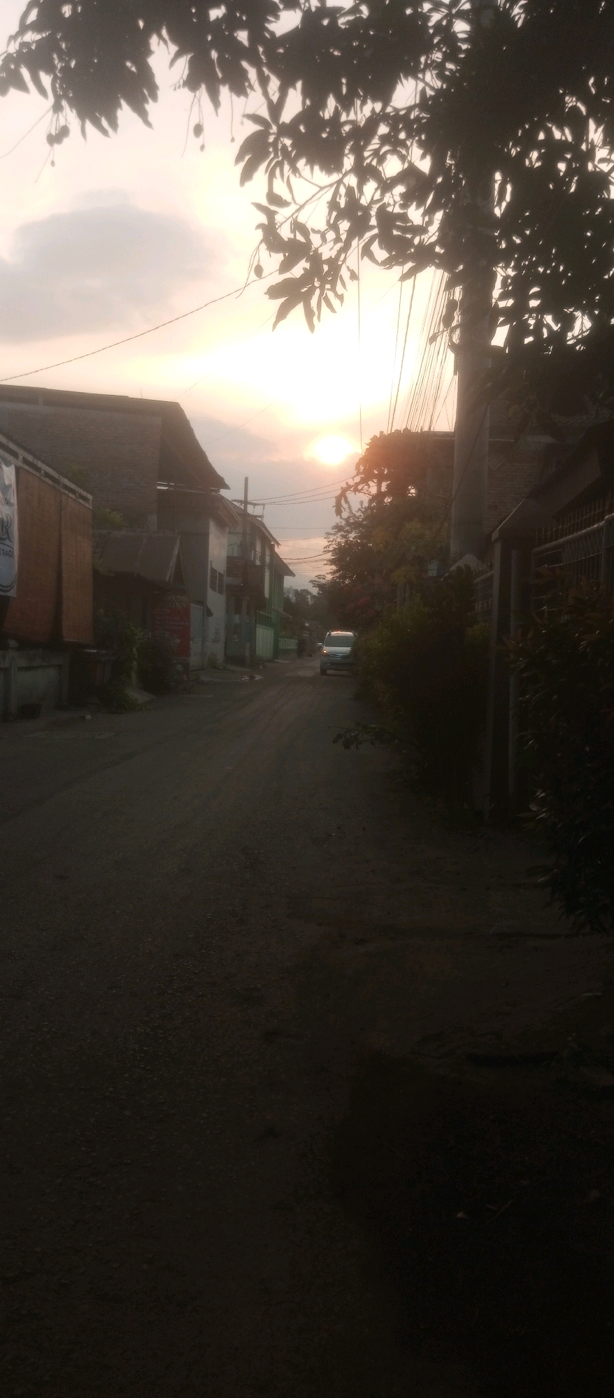 The image captures a residential street in Kediri City, Indonesia, during what appears to be late afternoon or early evening. The sun is low on the horizon, casting a warm, diffused light and creating silhouettes of trees and structures. On the left side of the street, buildings with corrugated metal roofs and bamboo-like awnings are visible. A white banner with some text is partially obscured. Further down the street, there are more buildings, some with multiple stories, painted in various colors including white, grey, and green.
A silver-colored vehicle, possibly a minivan or van, is parked or driving down the middle of the asphalt road. The road surface is textured and appears somewhat worn. On the right side of the street, dense foliage and trees obscure much of the residential buildings, with a fence or gate also present. Electrical wires crisscross overhead, contributing to the urban atmosphere. The sky is partly cloudy, with the sun creating a bright, hazy glow. The overall impression is of a quiet, lived-in neighborhood at the end of the day. There are no distinct individuals or clear actions depicted, suggesting a static moment captured in time.