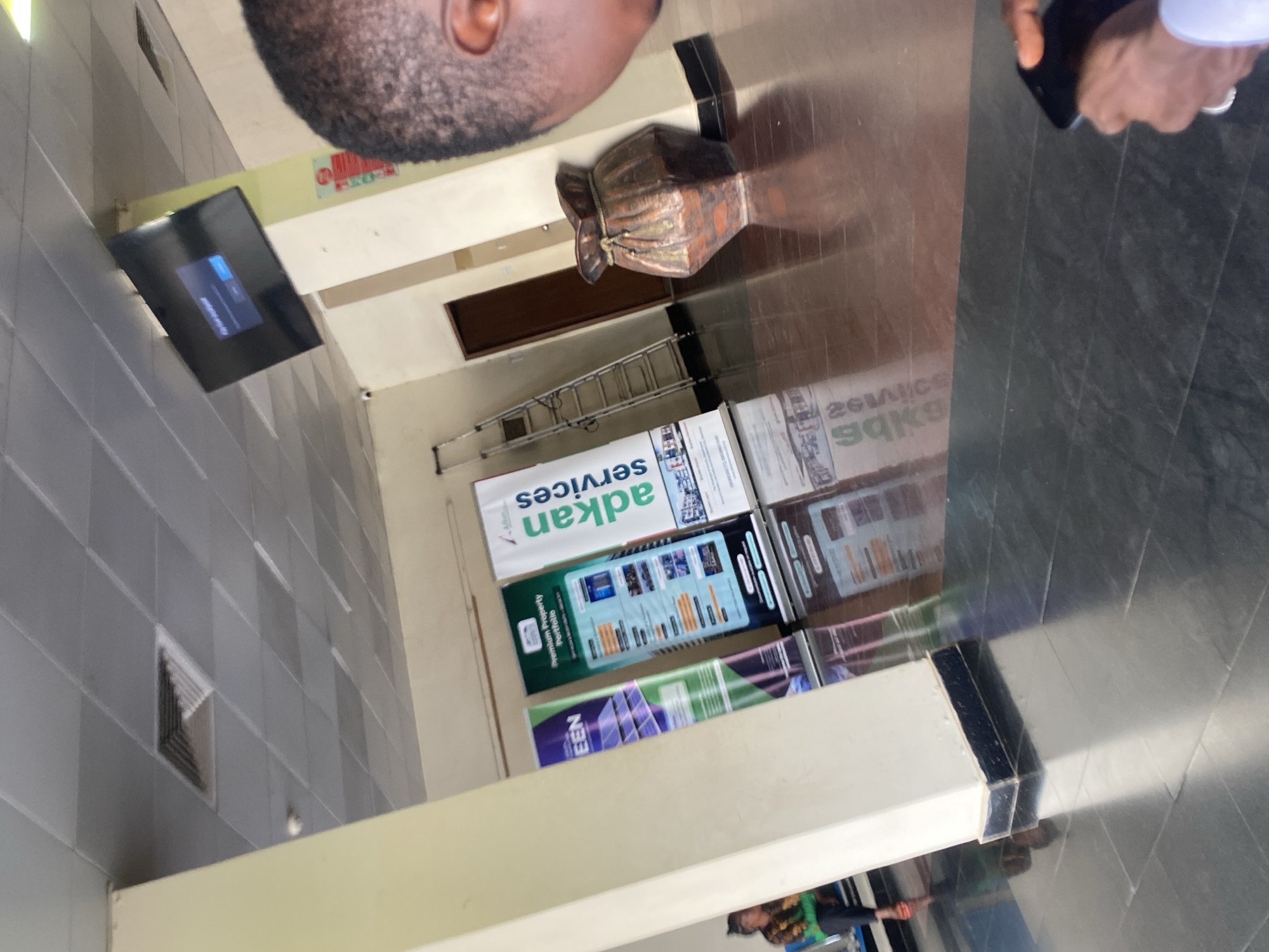 The image is taken from an unusual perspective, showing the upper part of a man's head and shoulder. The scene appears to be an indoor space, possibly a commercial area or a mall. A television screen is mounted on a wall. There are advertisements promoting "Adkan Services" and another displaying images of properties. A ladder leans against a wall near the advertisements. The floor is tiled and reflective. The location is Abuja, Nigeria, as indicated in the "Adkan Services" advertisement, which likely refers to a real estate or property management company operating in that city.
