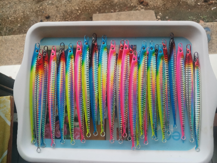 The image shows a white plastic tray filled with 21 fishing lures. The lures are arranged vertically in a row. They are all made of metal and have a silver base with colorful stripes of pink, blue, yellow, and green. Each lure has a small, sharp hook at the end and a loop at the other end. The lures are arranged in a neat, organized fashion and appear to be new and unused. The background is a blurred out view of a tiled floor. The image was taken in Kebumen, Indonesia at the coordinates -7.662, 109.647. The image was taken indoors and shows a close-up of the tray.